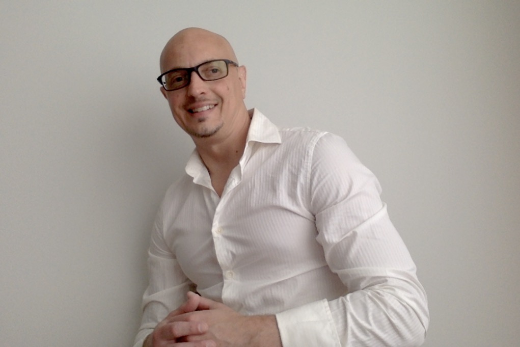 Man with glasses in a white shirt smiling, standing against a plain wall.