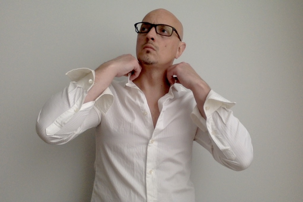 Bald man wearing glasses and a white shirt adjusting collar against plain wall.