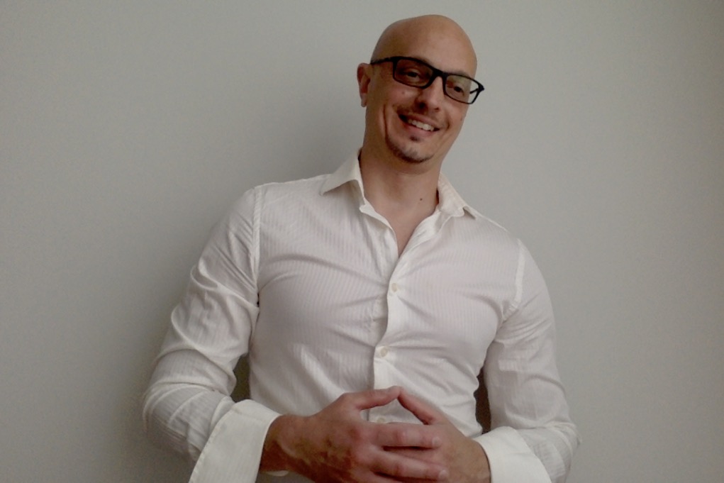 Bald man with glasses wearing a white shirt, smiling, standing against a plain wall.