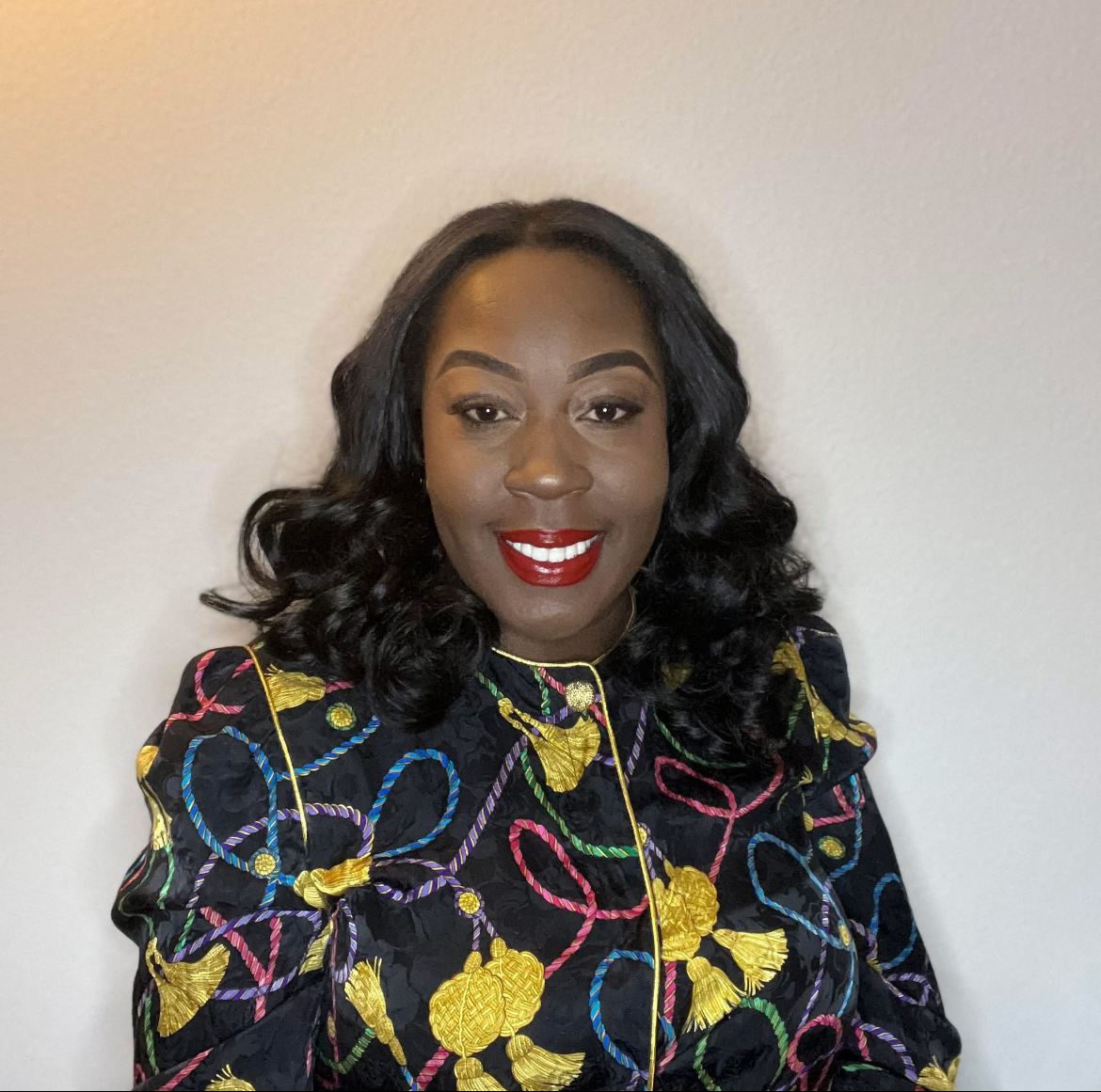 Woman with dark, wavy hair and red lipstick smiling, wearing a colorful patterned top, against a plain background.