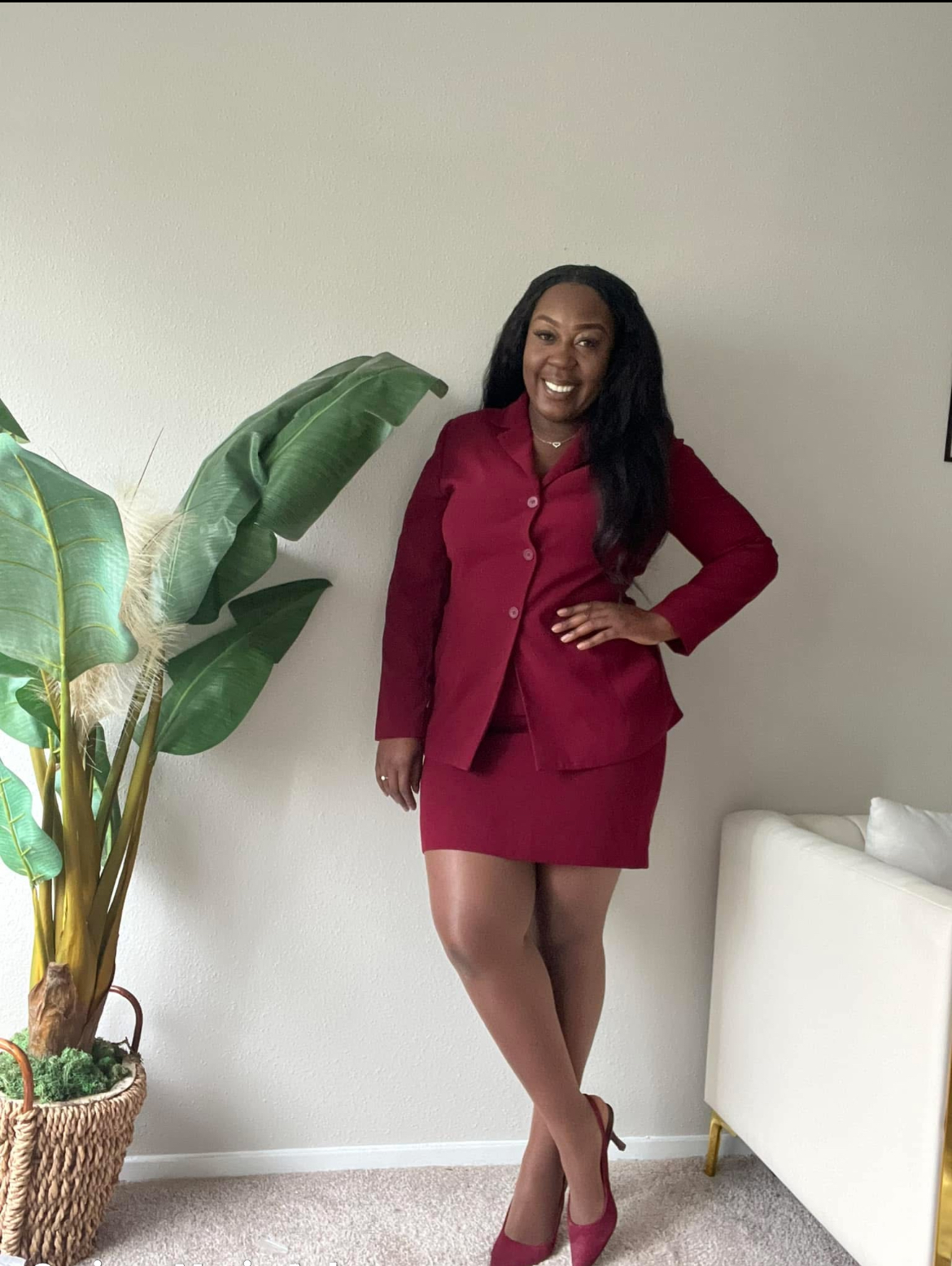 Woman in red outfit standing next to a large plant and a white sofa, smiling, with a plain wall background.