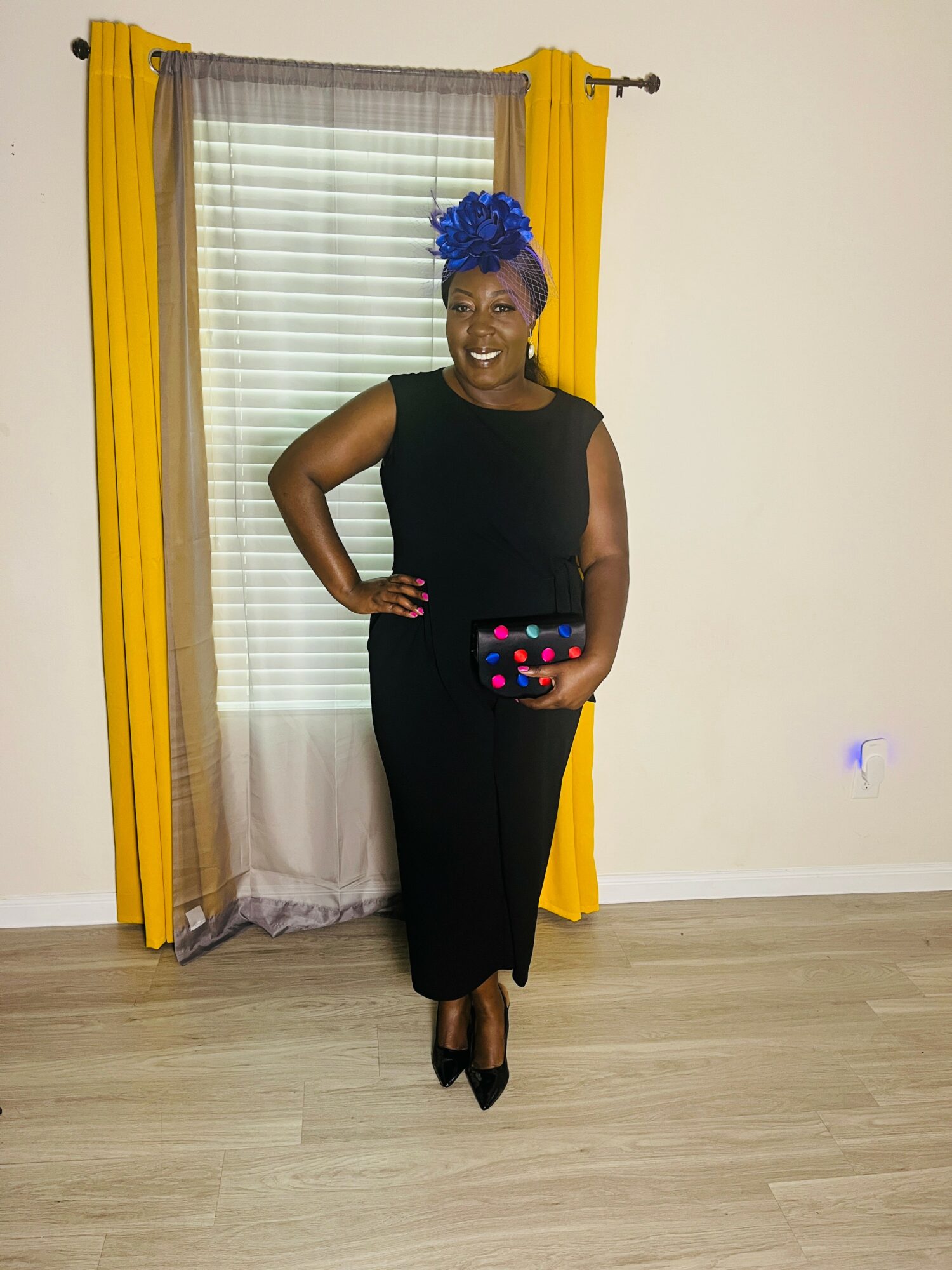 Woman in black dress with blue headwrap standing indoors, smiling, holding a colorful clutch, yellow curtains behind her.