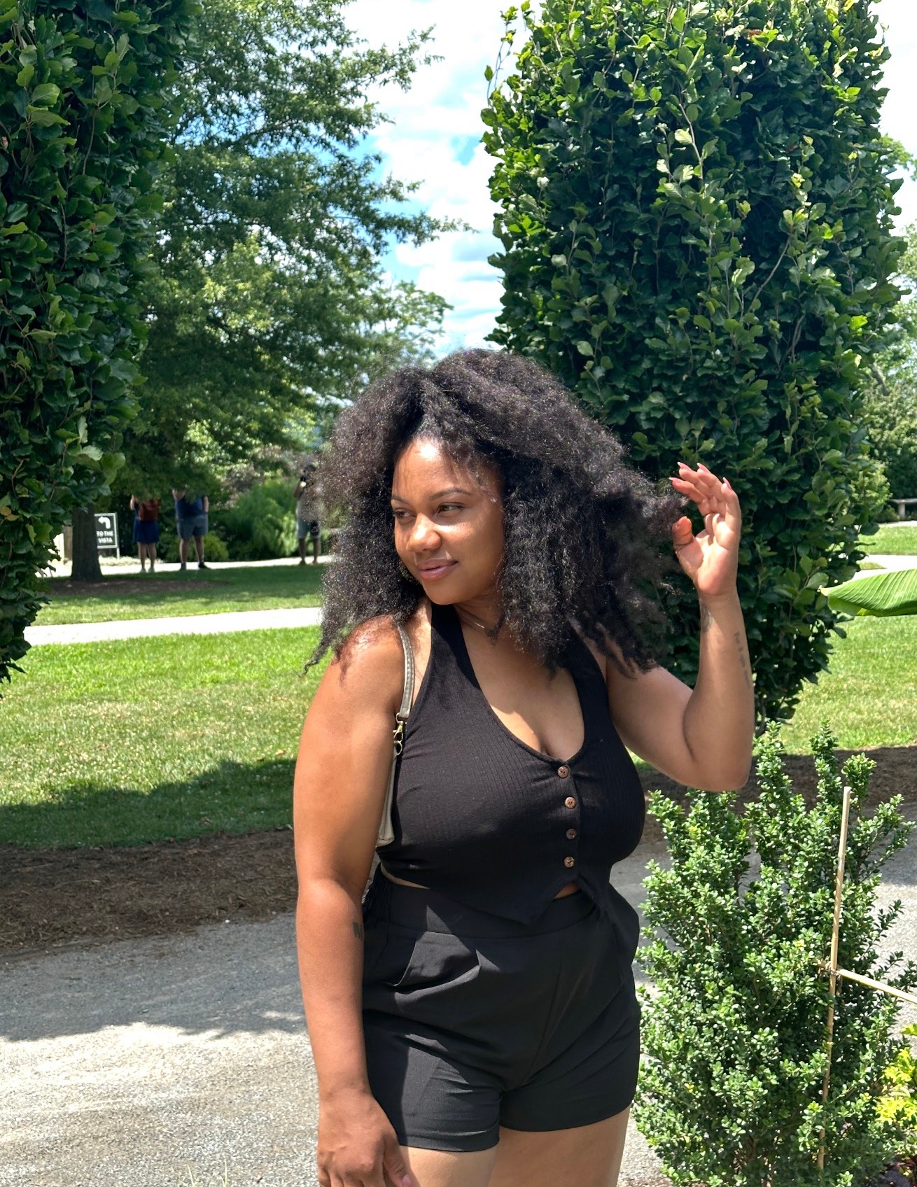 Young woman with curly hair standing outdoors in a park, smiling, wearing a black sleeveless top and shorts, trees in background.