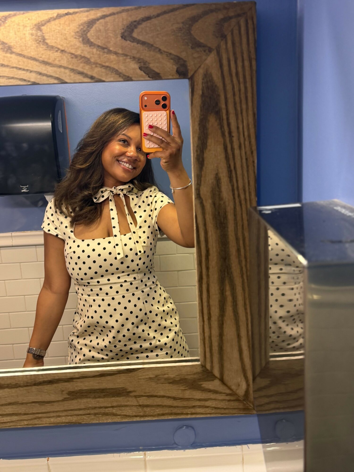 Woman taking a mirror selfie with a pink phone, wearing a polka dot dress, in a room with wooden and blue walls.