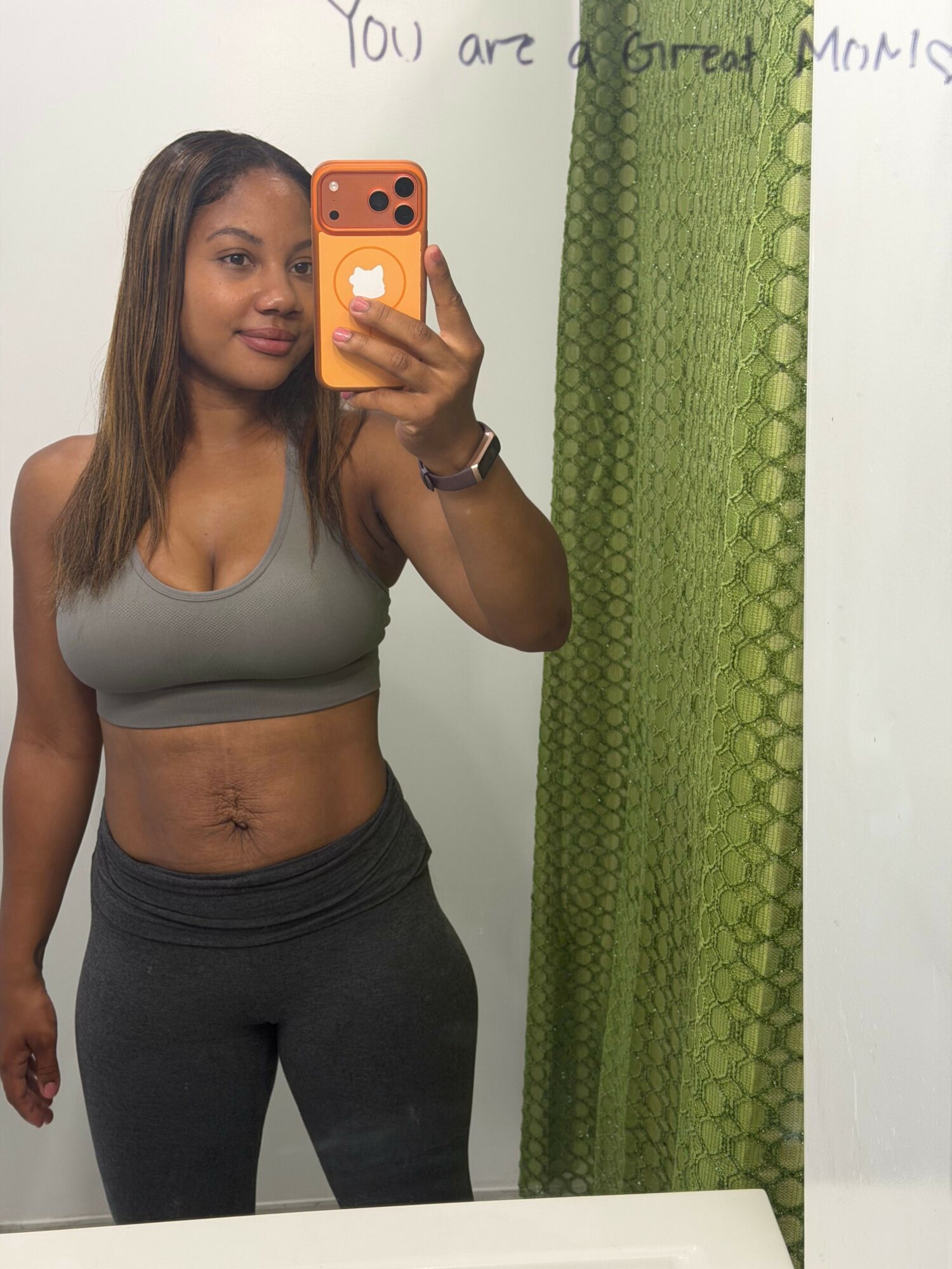 Woman taking a mirror selfie with a smartphone, wearing a gray top and black pants, standing in front of a green curtain.