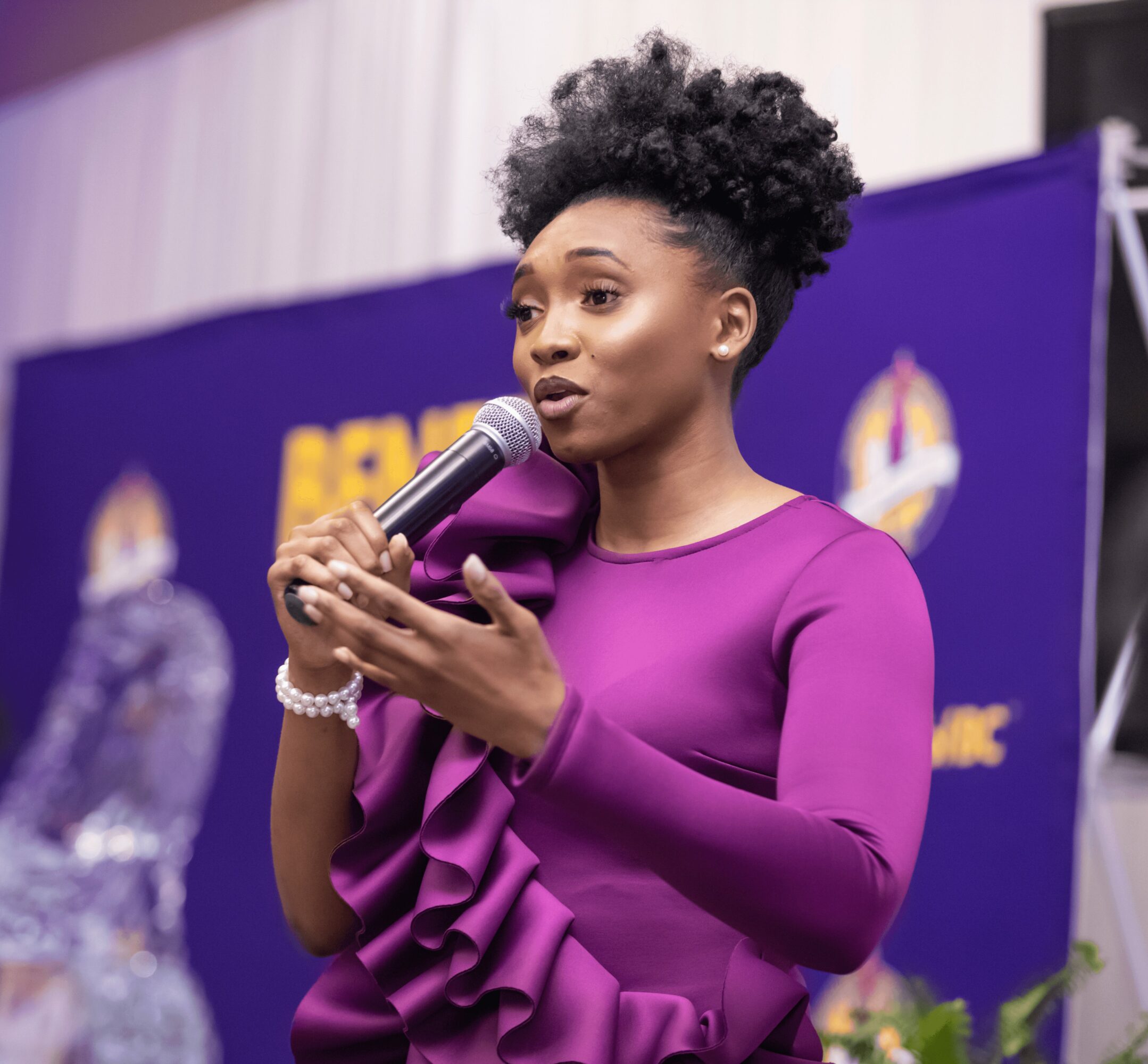 Woman with natural hair speaking into a microphone, wearing a purple dress with ruffles, in front of a blue backdrop.