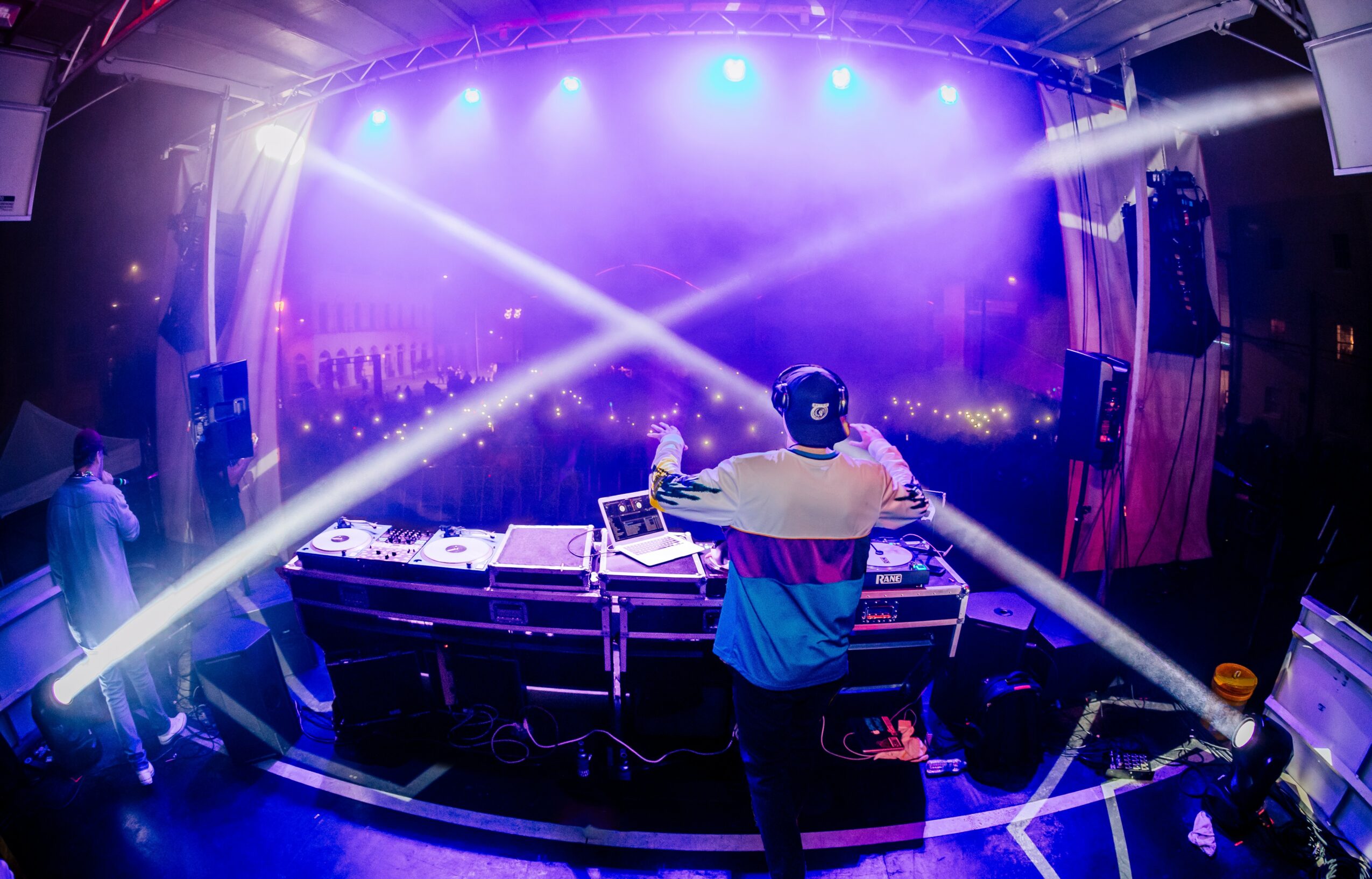 DJ performs on stage with colorful lights and fog, facing a crowd at a concert or event, with equipment in front.