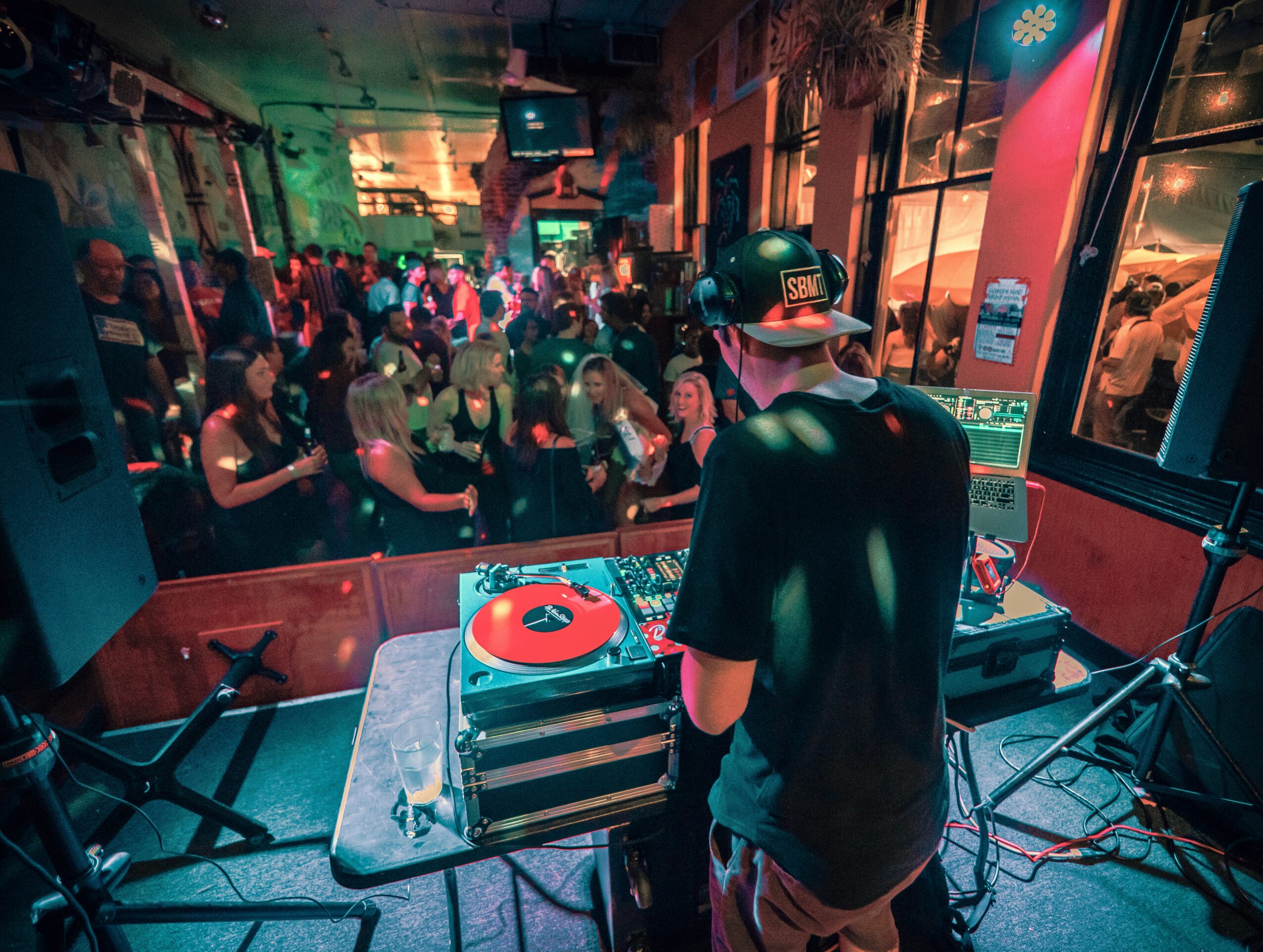 DJ wearing a cap and headphones performs at a crowded indoor dance event with colorful lighting.