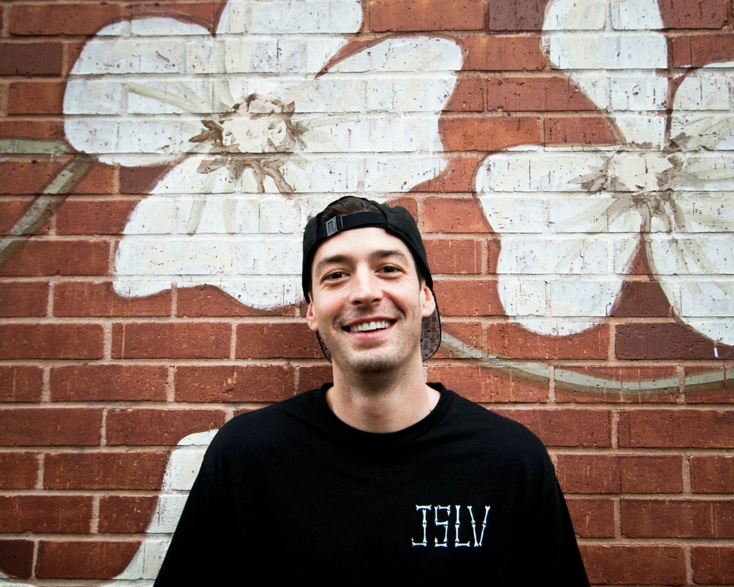 Young man with a black cap and shirt smiling in front of a brick wall with white flower mural.