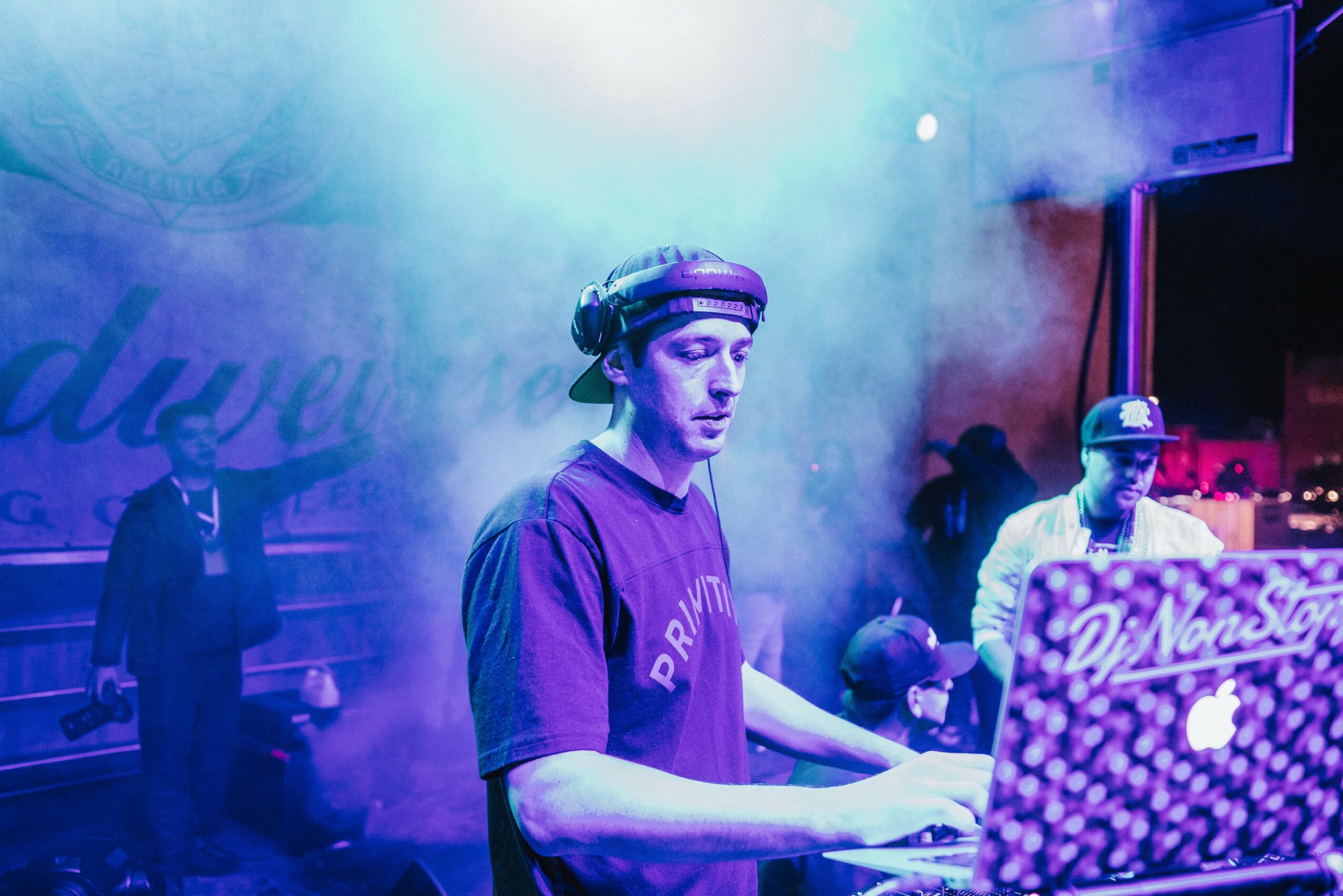 DJ wearing headphones operates equipment at a party with colorful lighting and smoke effects, people in the background. Word 'Dew' visible.