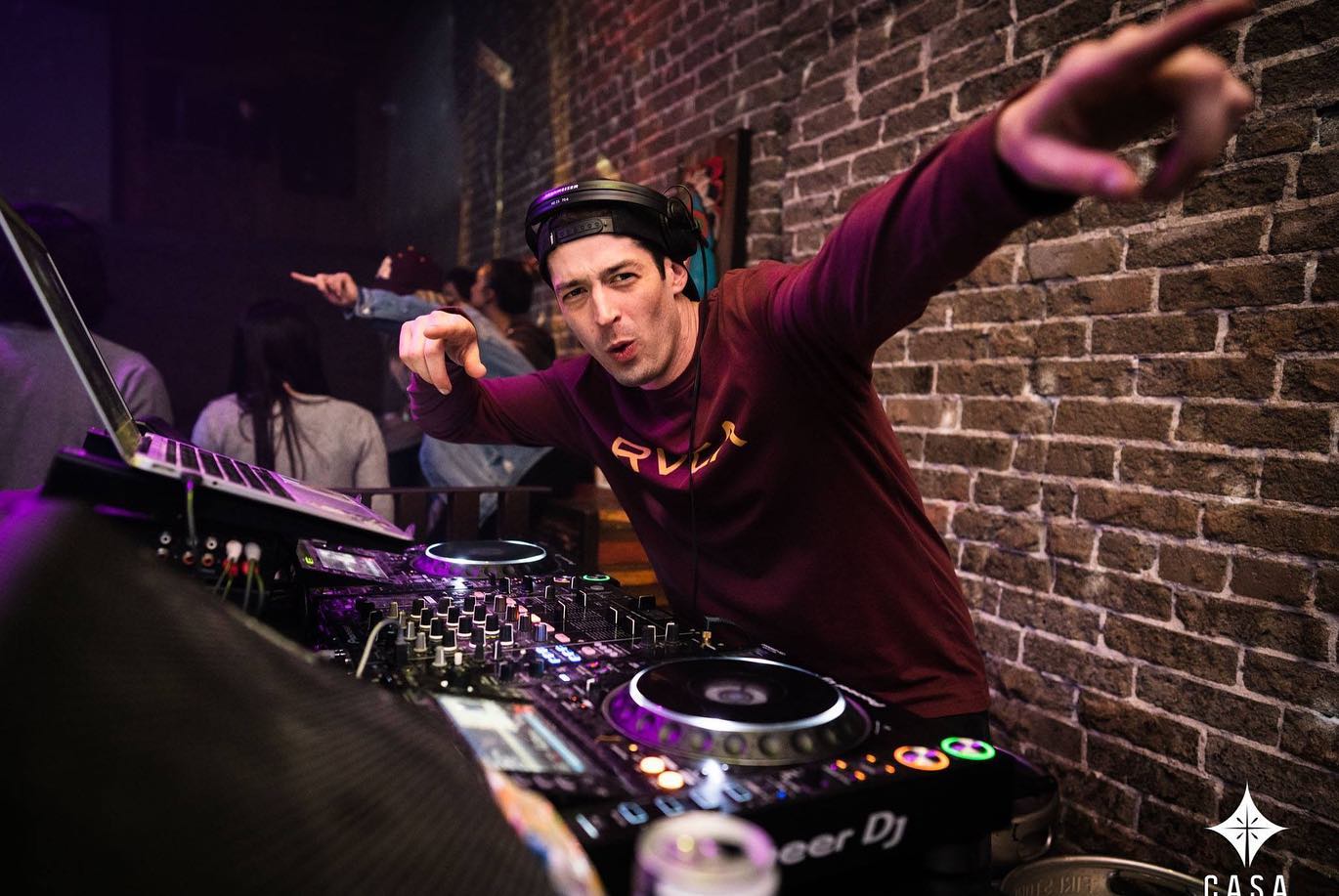 DJ wearing headphones points towards the camera in a brick-walled venue, with people in the background.
