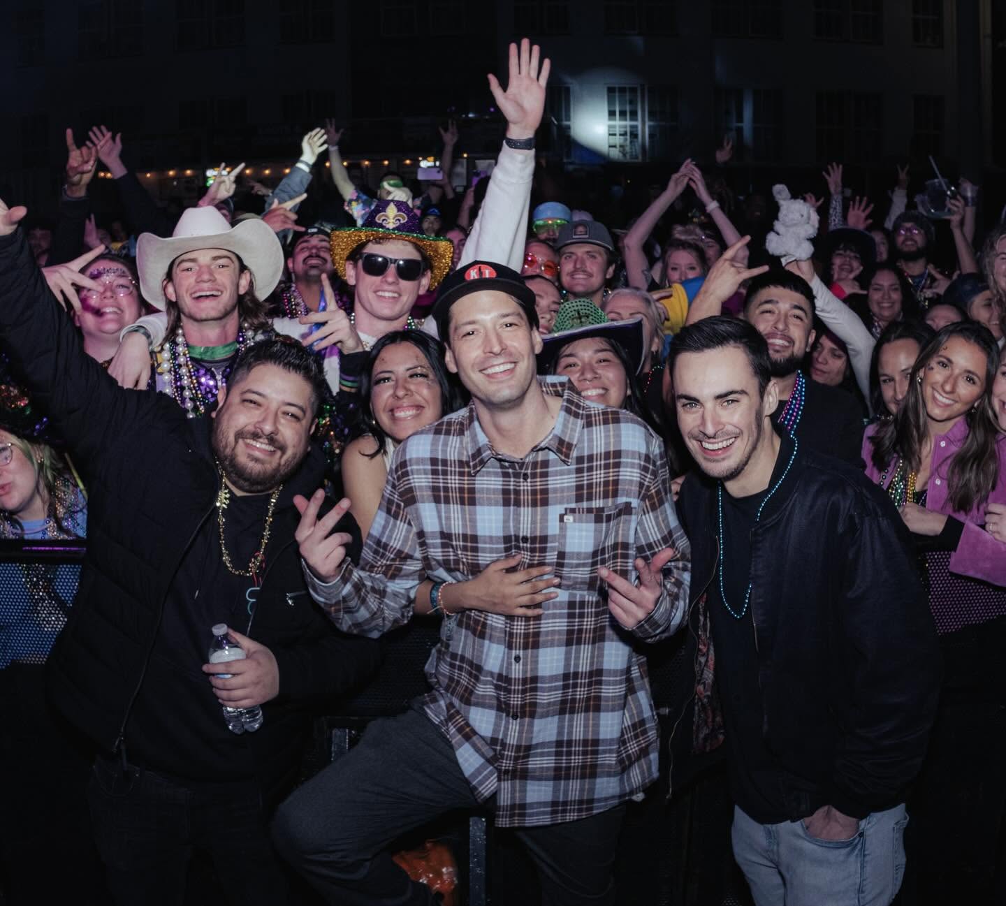 Group of people at a celebration, smiling and raising hands, with some wearing hats and beads, outdoors at night.
