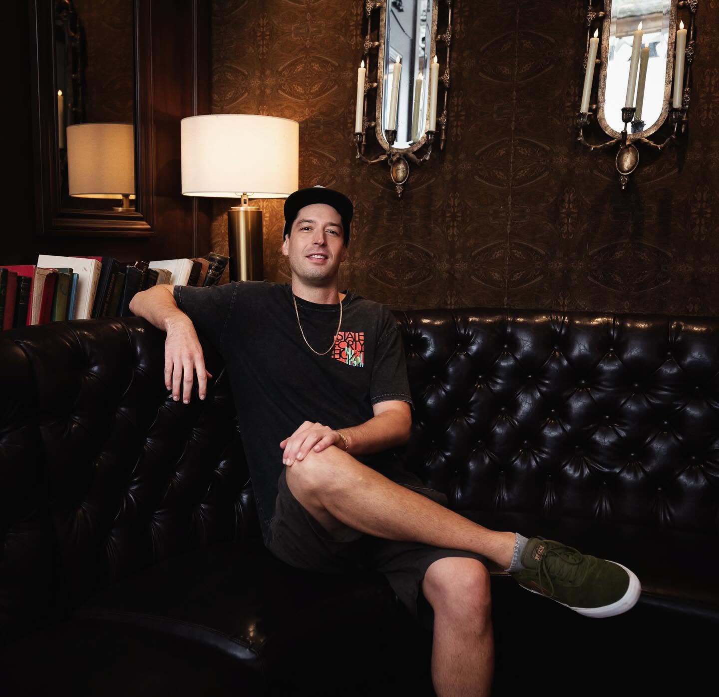 Young man sitting on a black leather couch in a dimly lit room with mirrors and lamps, smiling, with crossed legs.