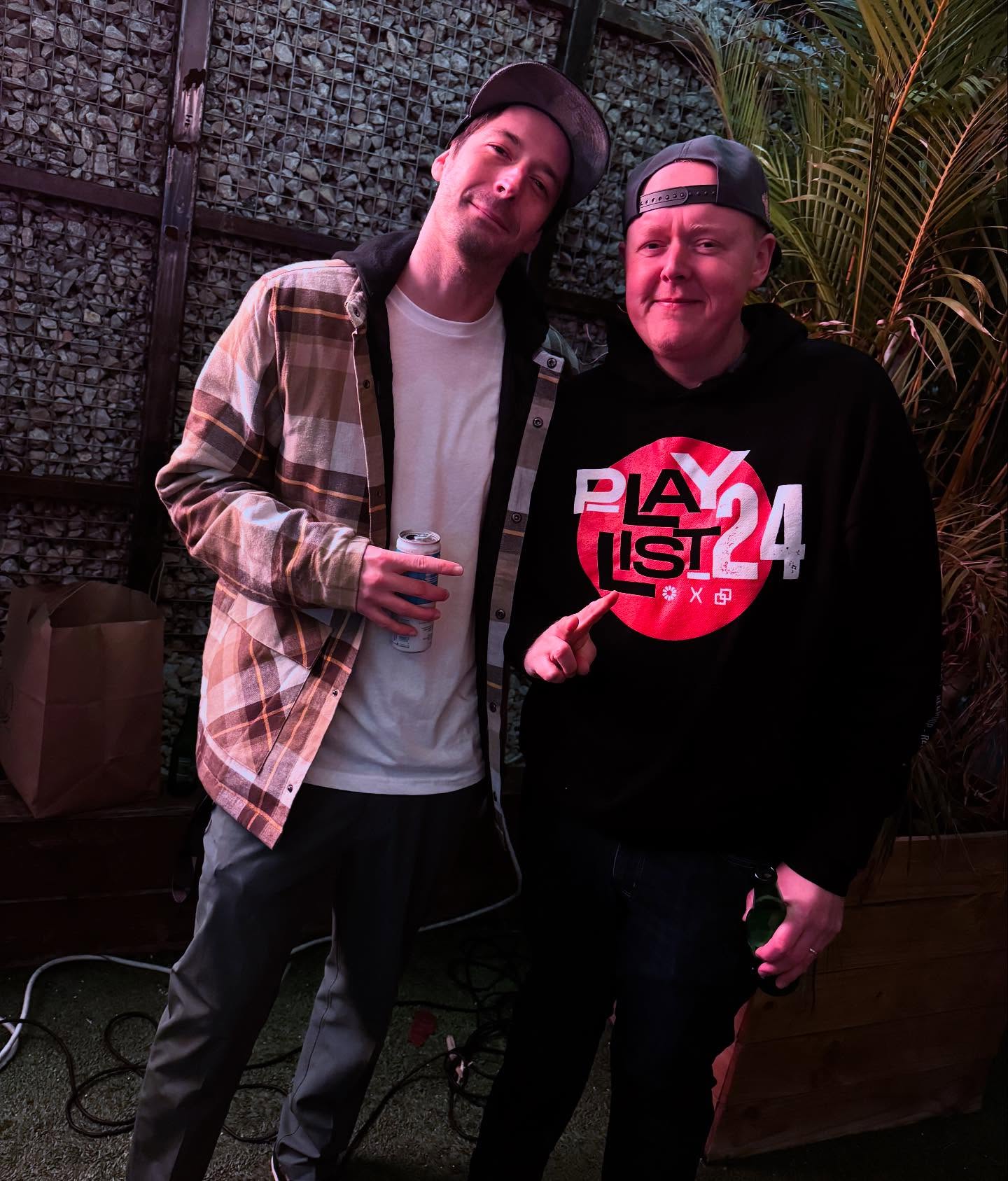 Two men standing outdoors, one holding a drink, with plants and a textured wall behind them.