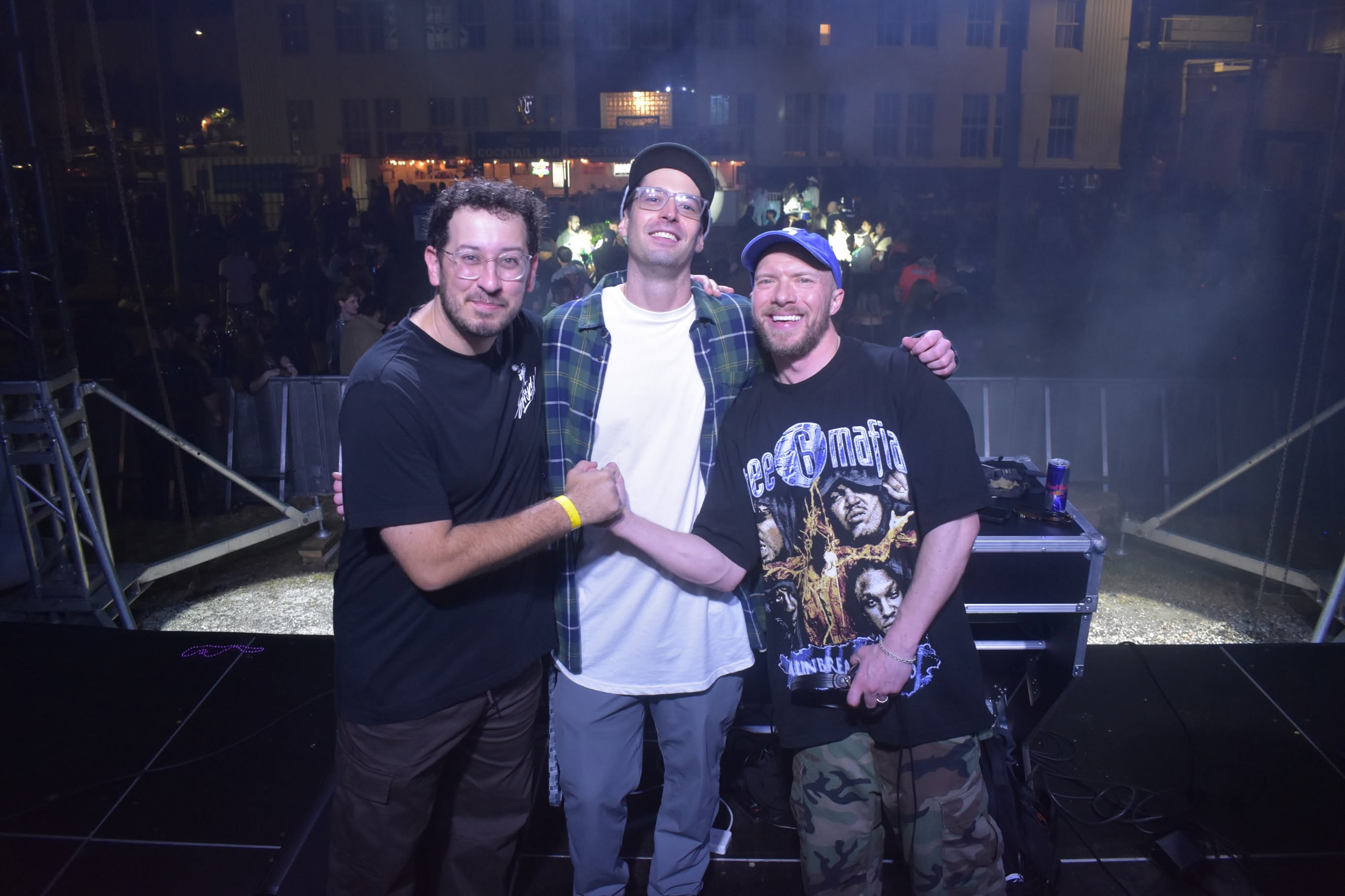 Three men standing together on a stage at night, smiling, with a crowd and city lights in the background.