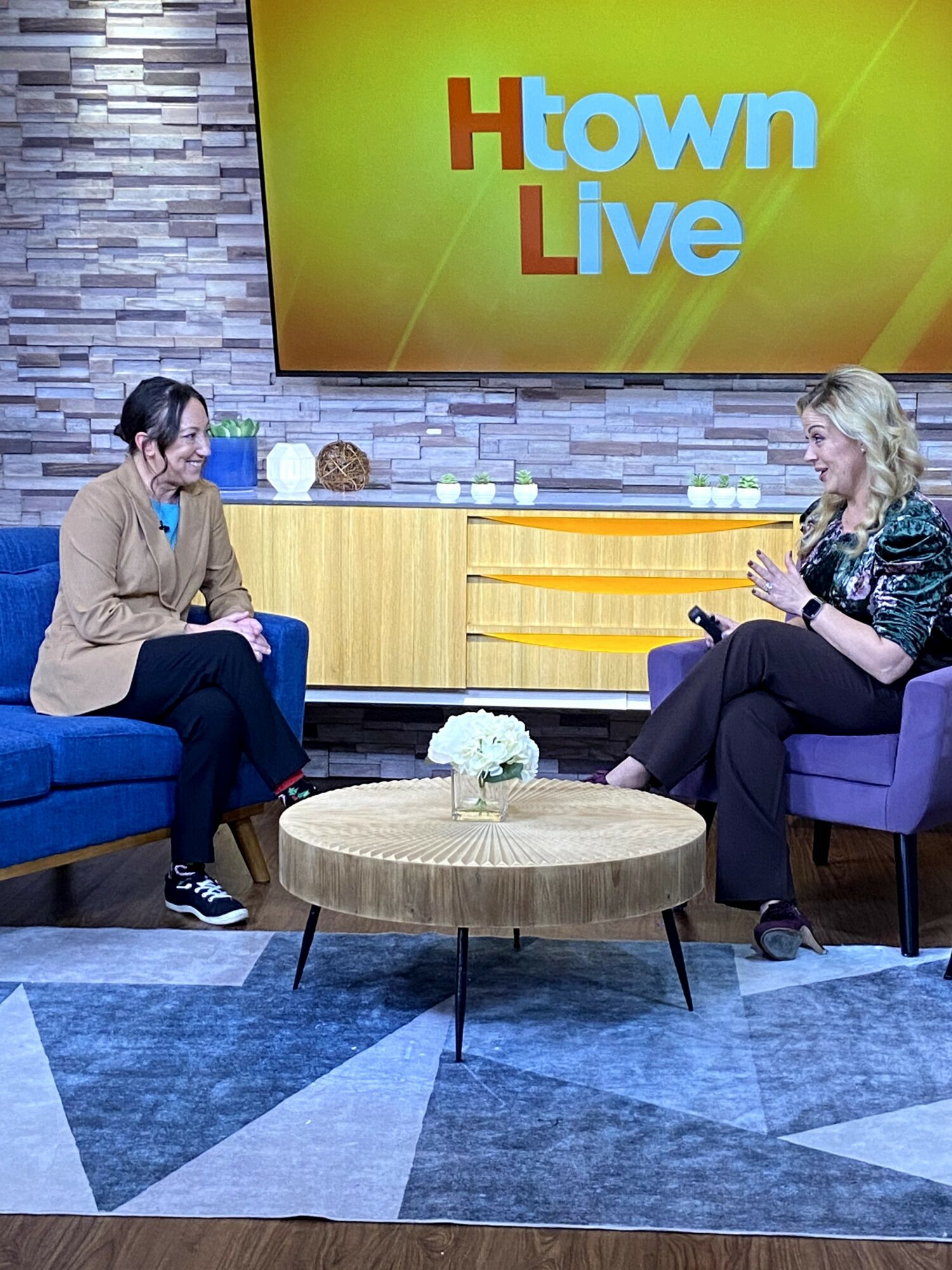 Two women sit on chairs facing each other in a TV studio with a yellow and gray background and a large screen displaying 'Htown Live'.