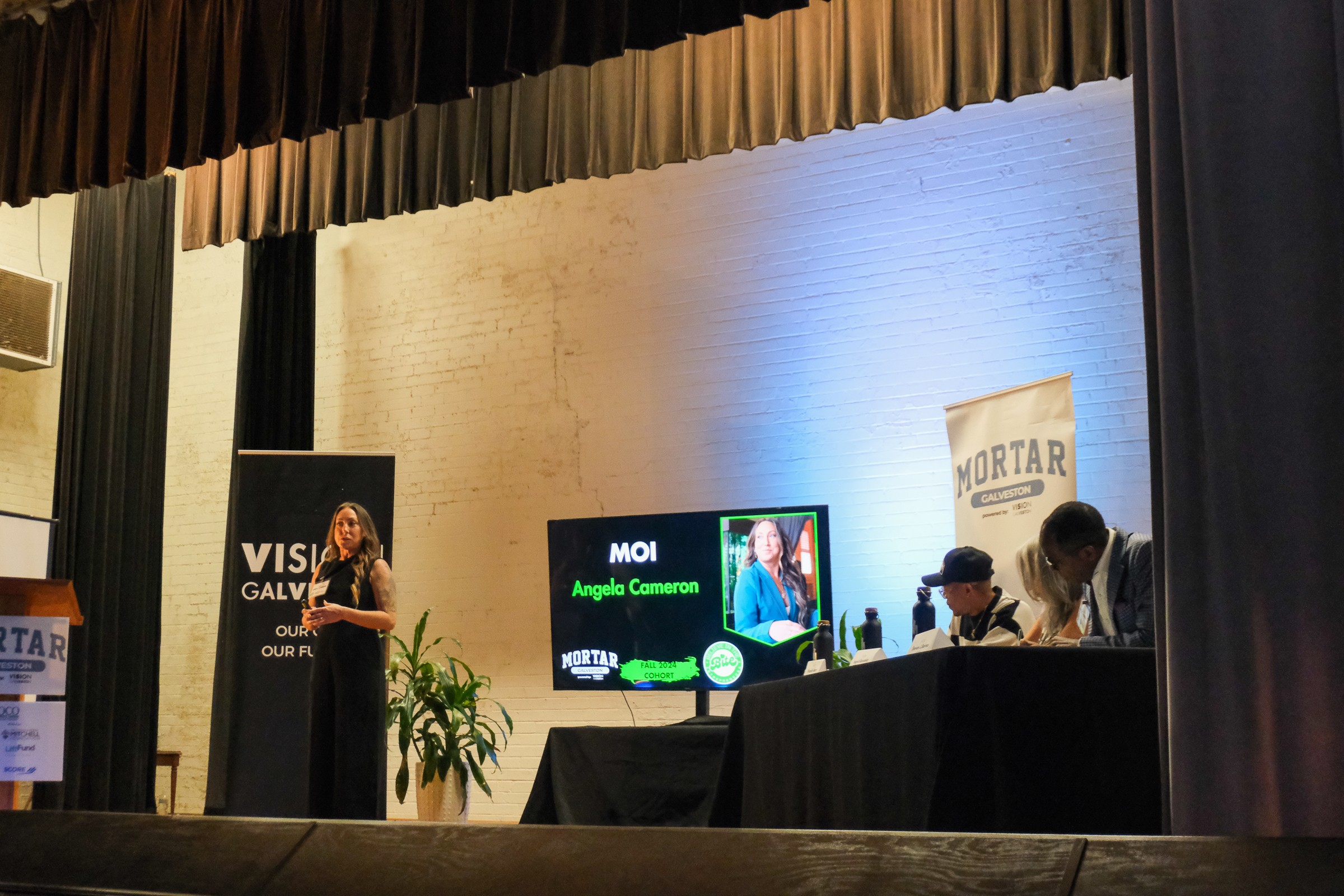A woman stands at a podium on stage, speaking to an audience, with a panel of three people seated at a table nearby.