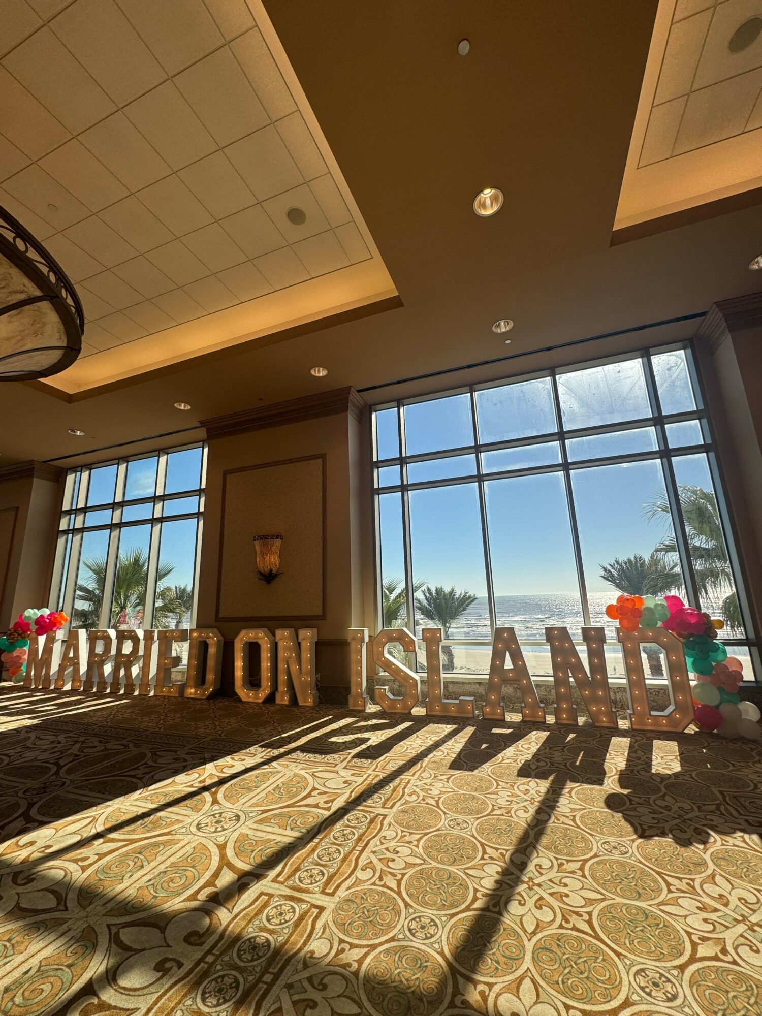 Indoor space with large windows showing outdoor palm trees, decorated with colorful balloons, and a sign reading 'WELCOME' and 'CONGRATULATIONS'.
