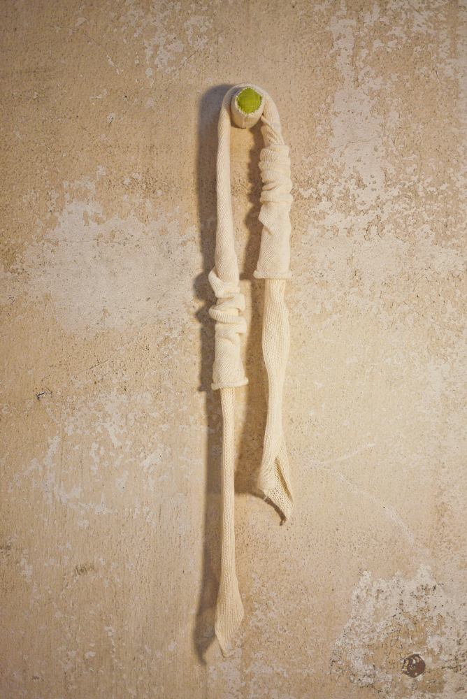A long, twisted object hanging vertically on a textured beige wall, with a small green and white item at the top.