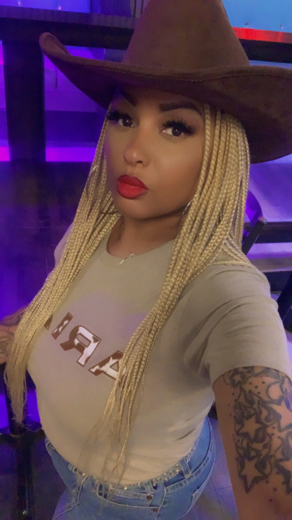 Woman with long braided hair wearing a large hat and red lipstick, taking a selfie indoors.