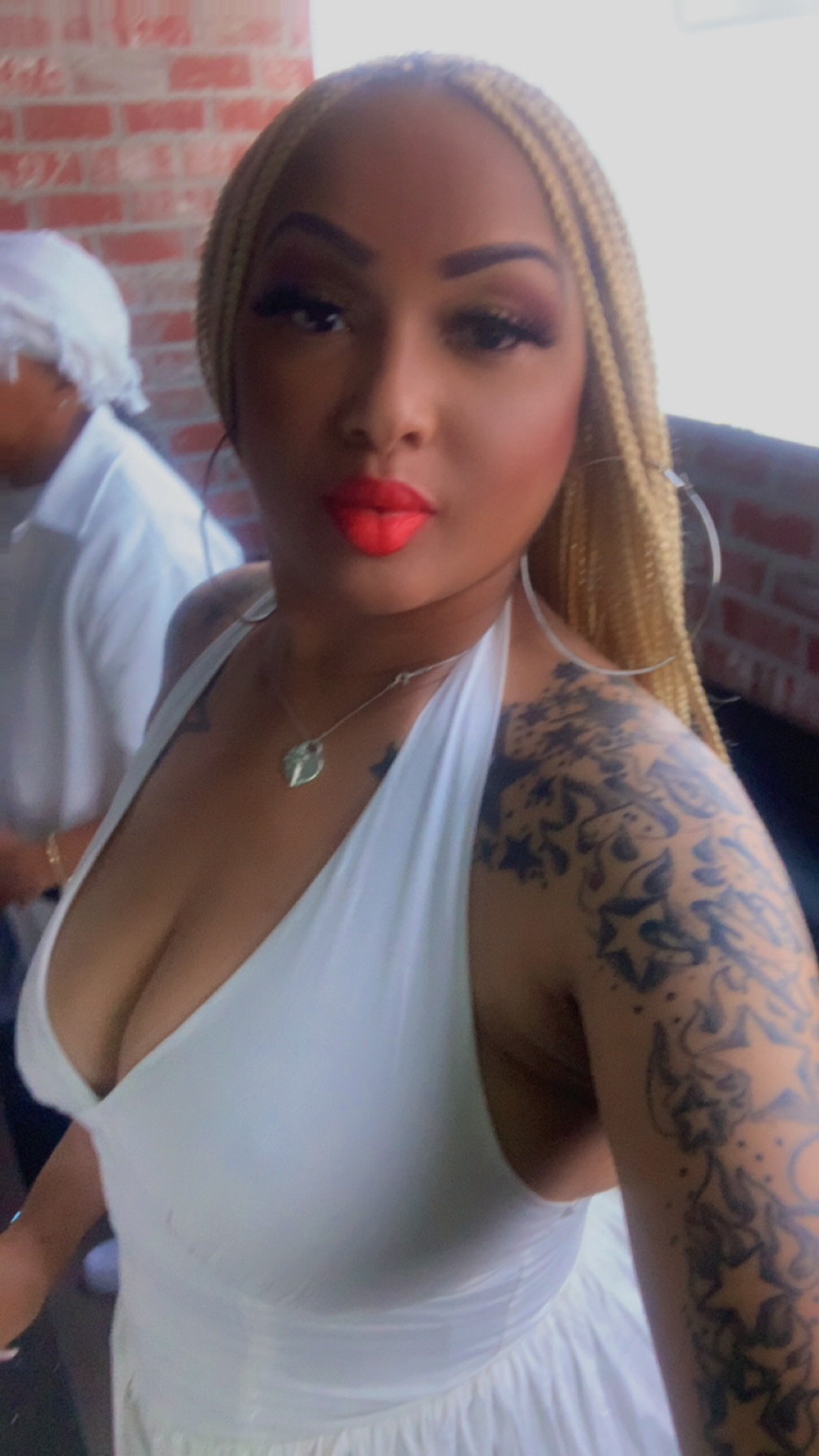 Woman with long blonde hair, red lipstick, wearing a white top, showing a tattooed arm, in front of a brick wall.
