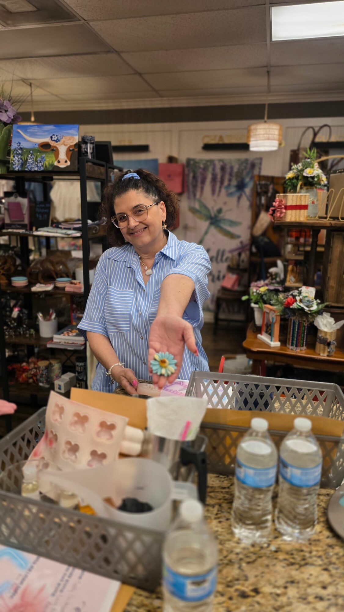 Woman in blue shirt holding a small object, standing in a store with shelves and various items around her.