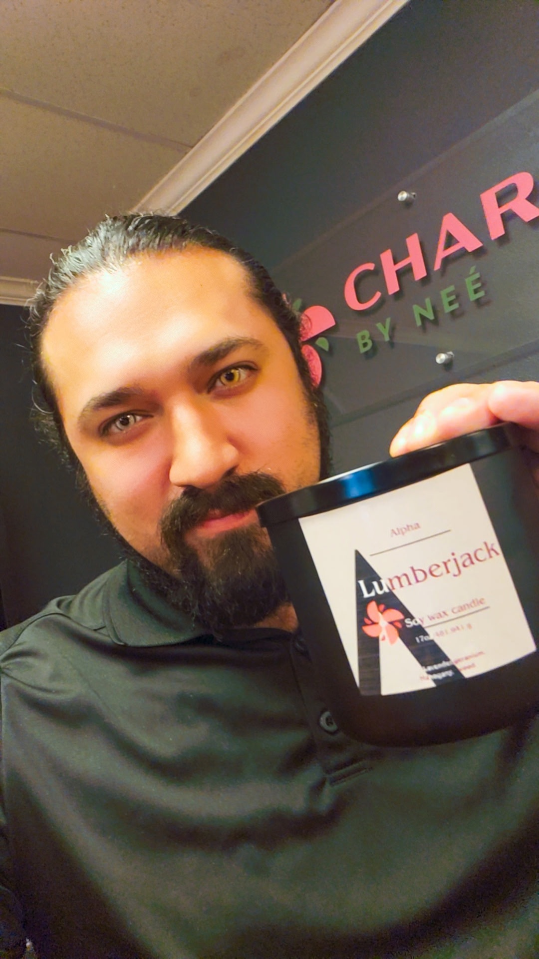 Man with dark hair and beard holding a container with a label, standing in front of a sign with pink and green text.