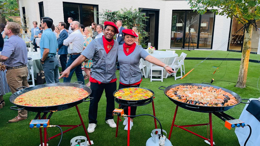 Two people in gray shirts and red bandanas stand behind three large pans of food outdoors, with a group of people in the background.