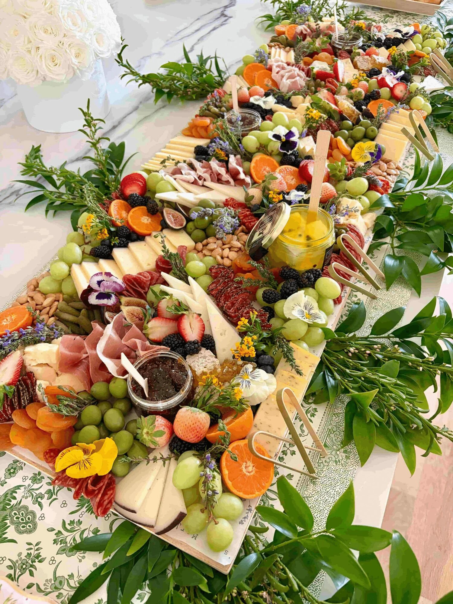 Colorful fruit and cheese platter with grapes, berries, and vegetables on a long wooden board, decorated with greenery.