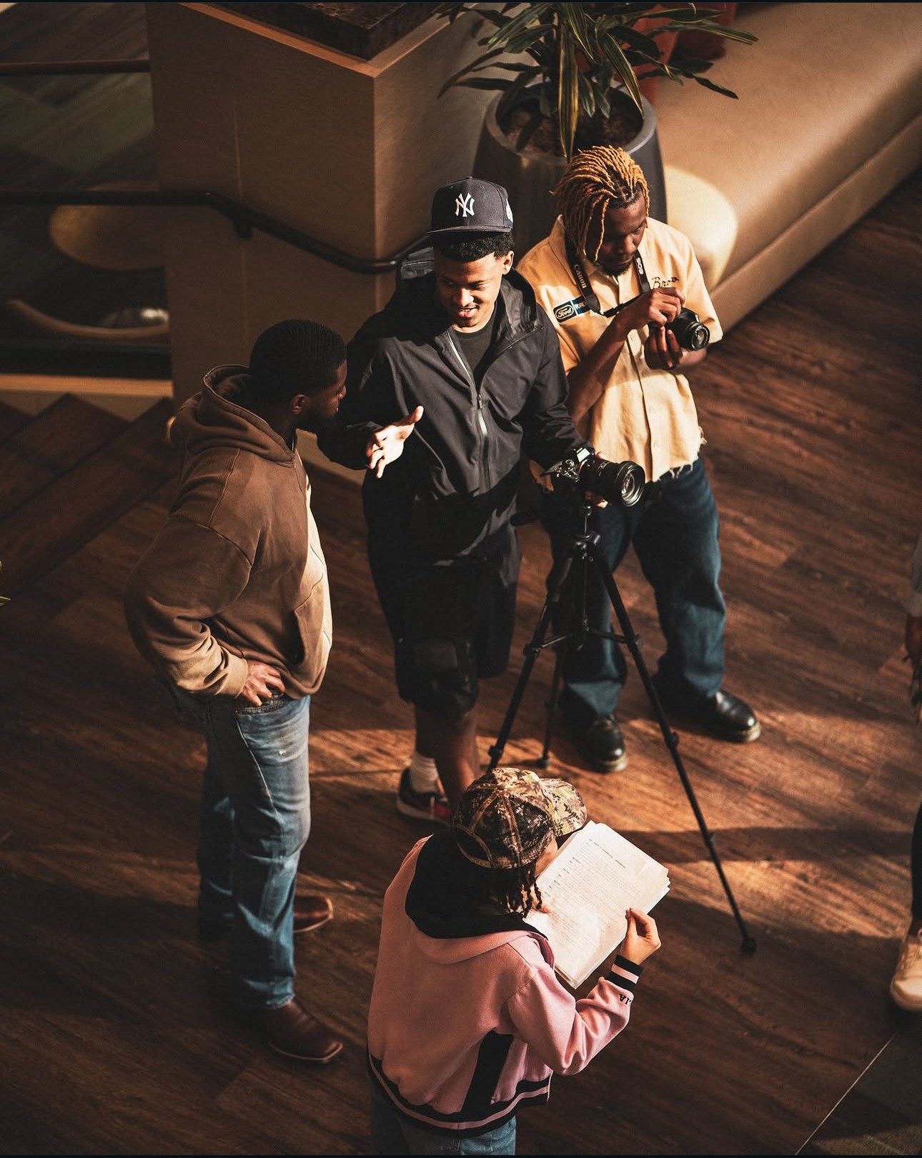 Four people stand on a wooden floor, one taking a photo with a camera on a tripod, others watching or writing.
