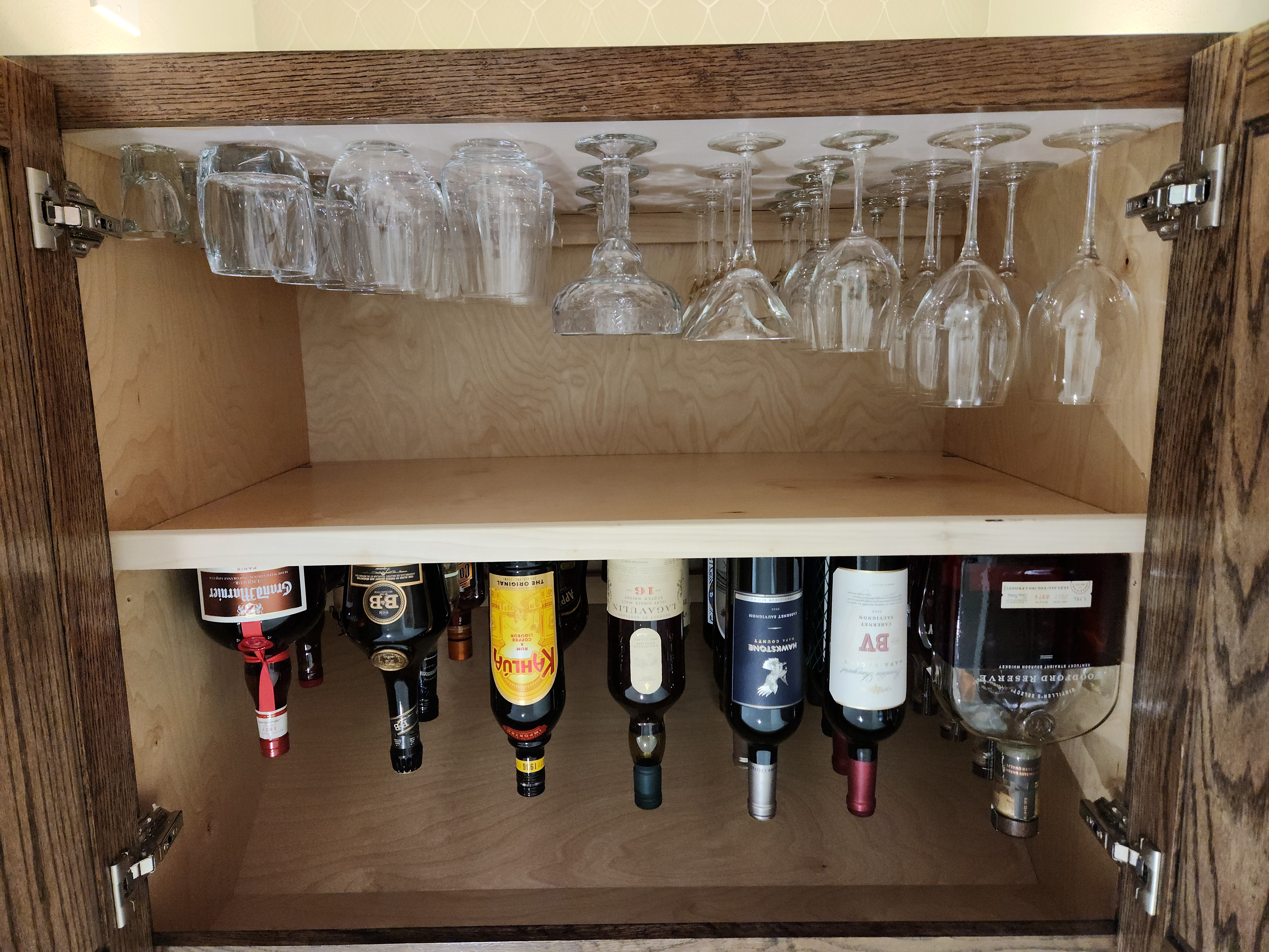 Open cabinet with hanging wine glasses above and bottles of alcohol on shelf below, against a beige wall.
