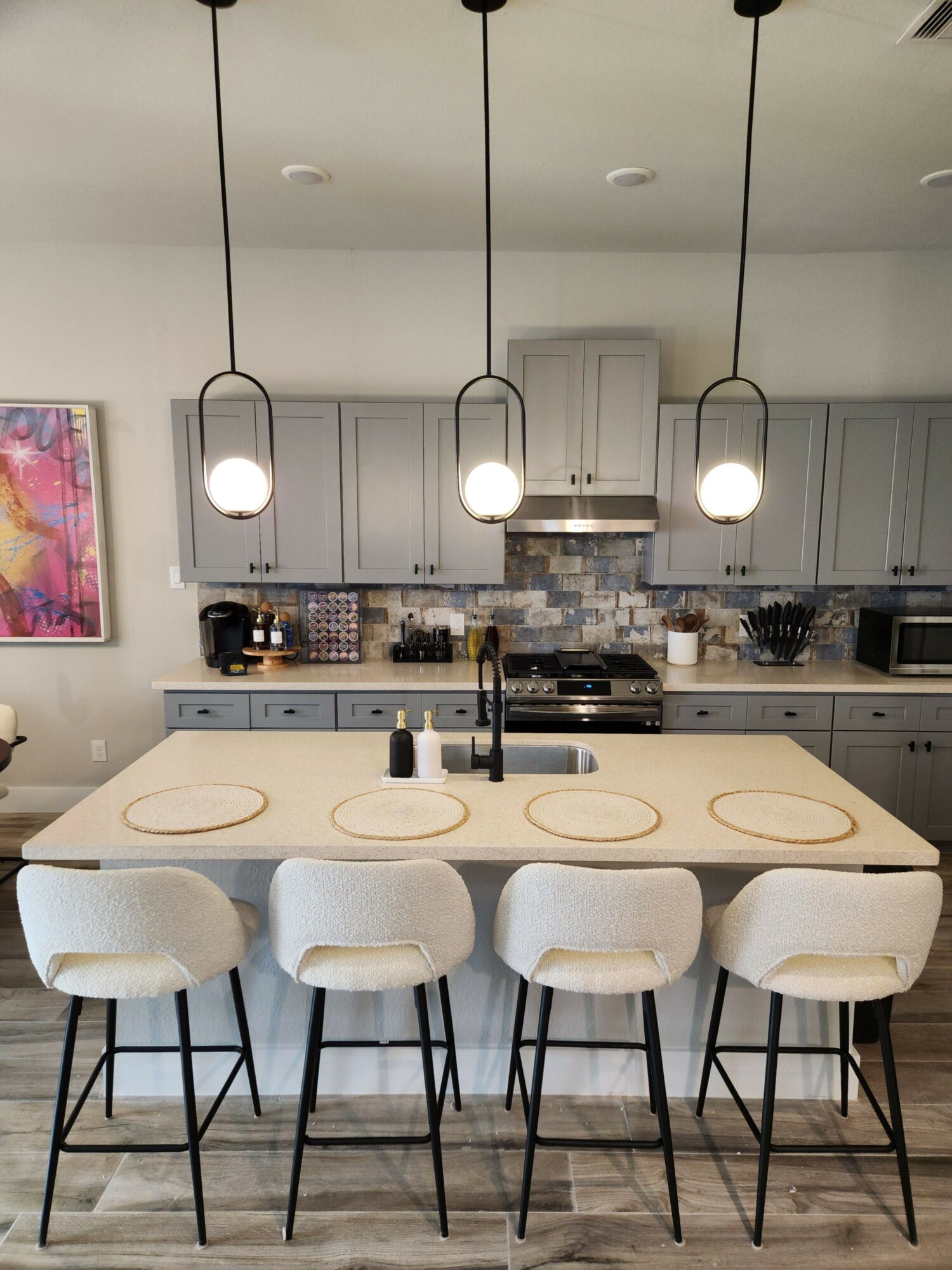 Dining area with a table and six white chairs, kitchen in background, pendant lights hanging, and a colorful artwork on the wall.