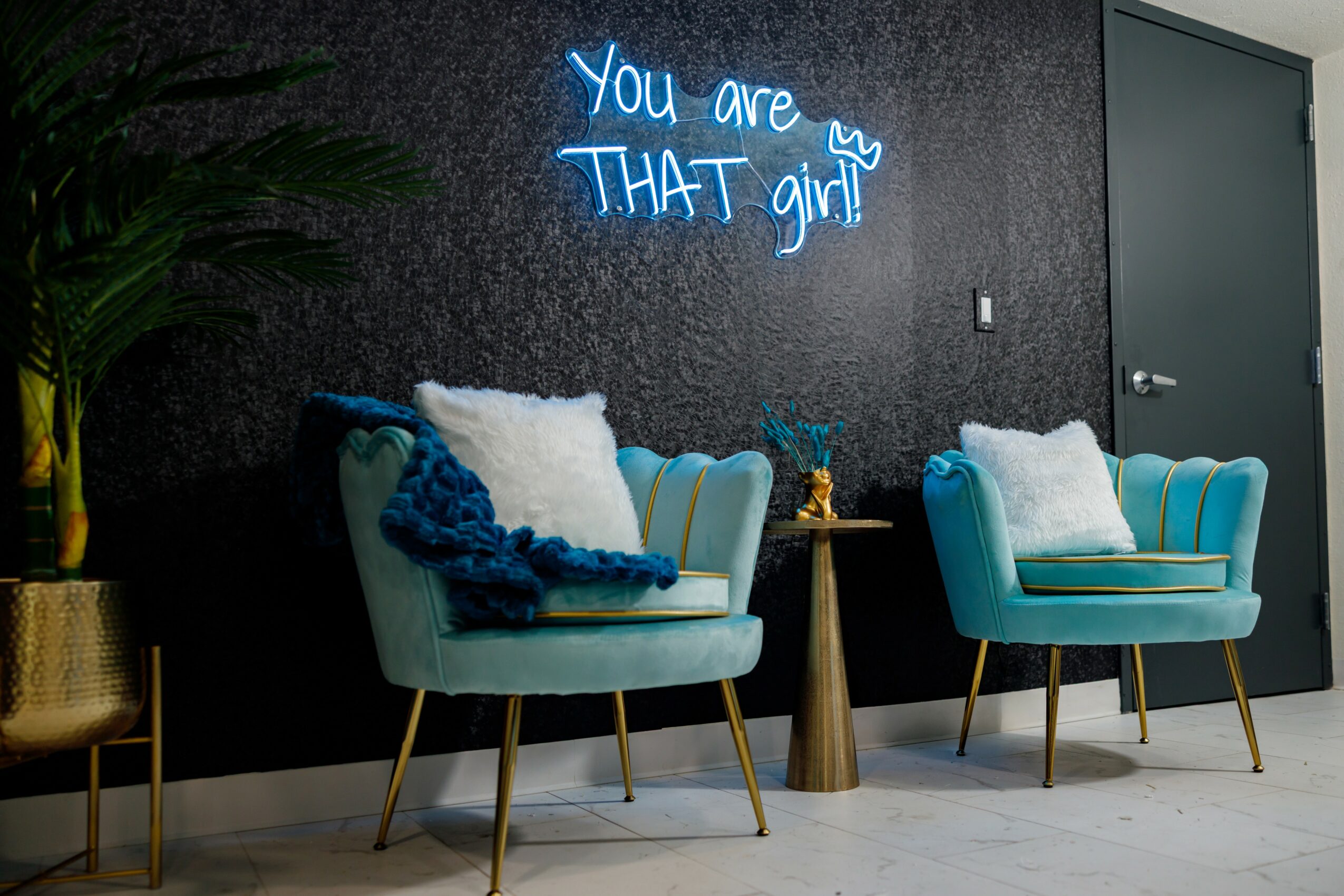 Two turquoise armchairs with white pillows and a blue throw, a small round table between them, and a neon sign on a dark wall.