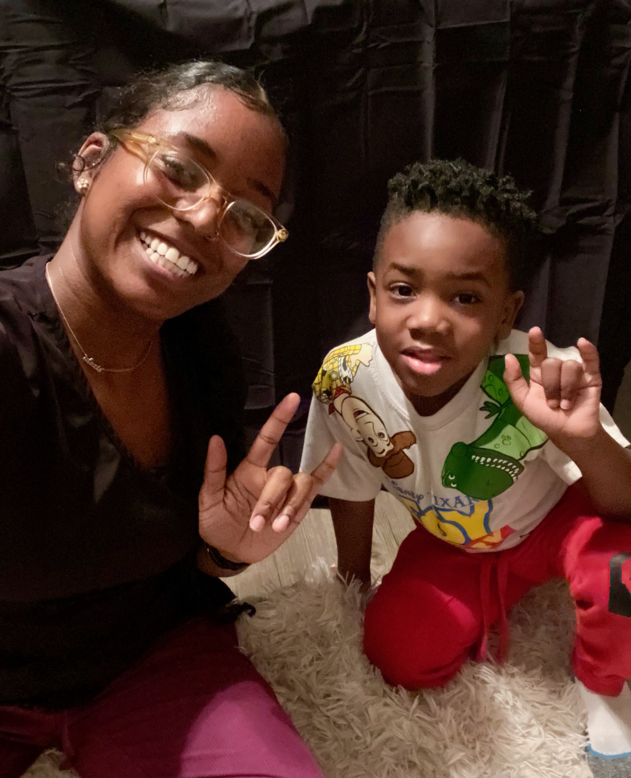 Smiling woman and young boy making hand gestures, sitting on a fluffy rug indoors.