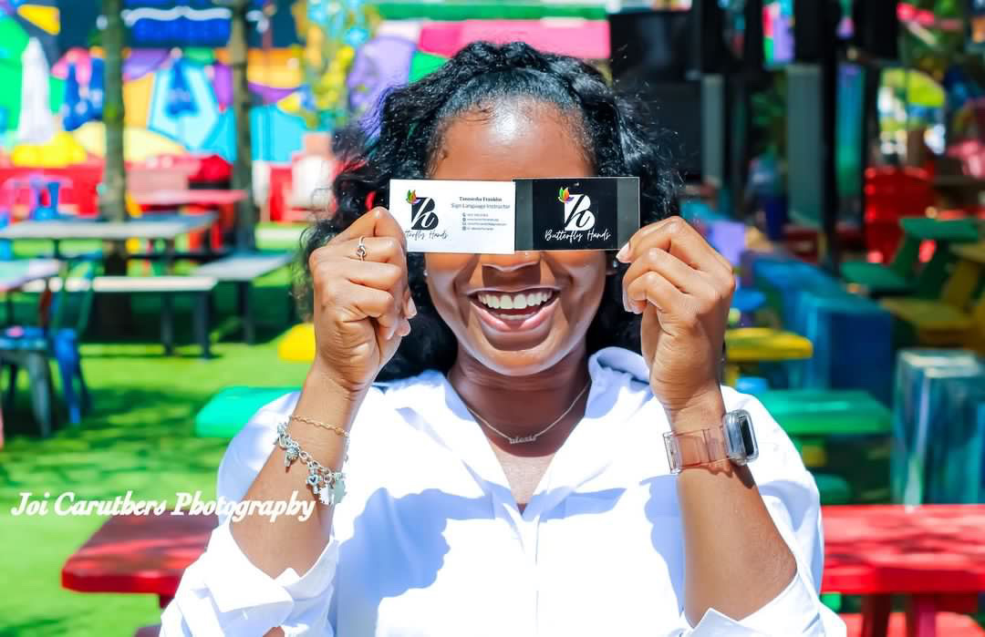 Woman holding a black and white card in front of her eyes, smiling, colorful outdoor setting with benches and trees