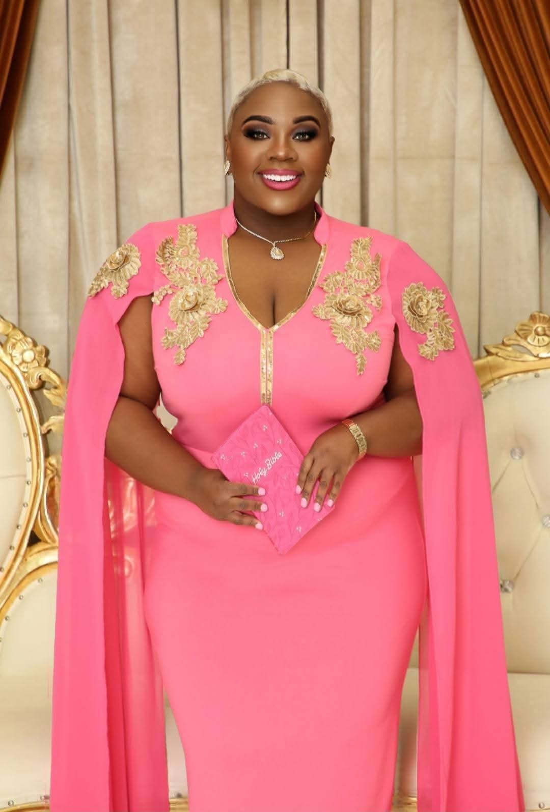 Woman in pink dress with gold embroidery, standing indoors with beige curtains and gold accents behind her.