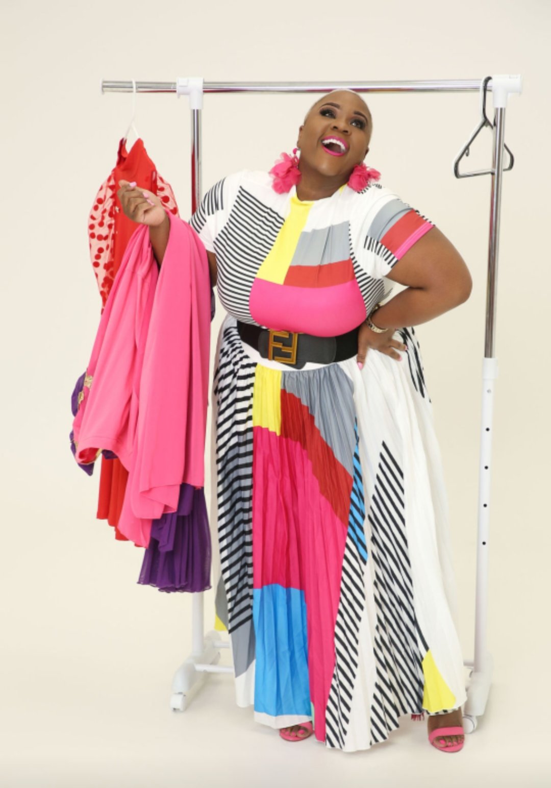 Person standing inside a clothing rack holding colorful dresses, wearing a multicolored striped dress, with a joyful expression.