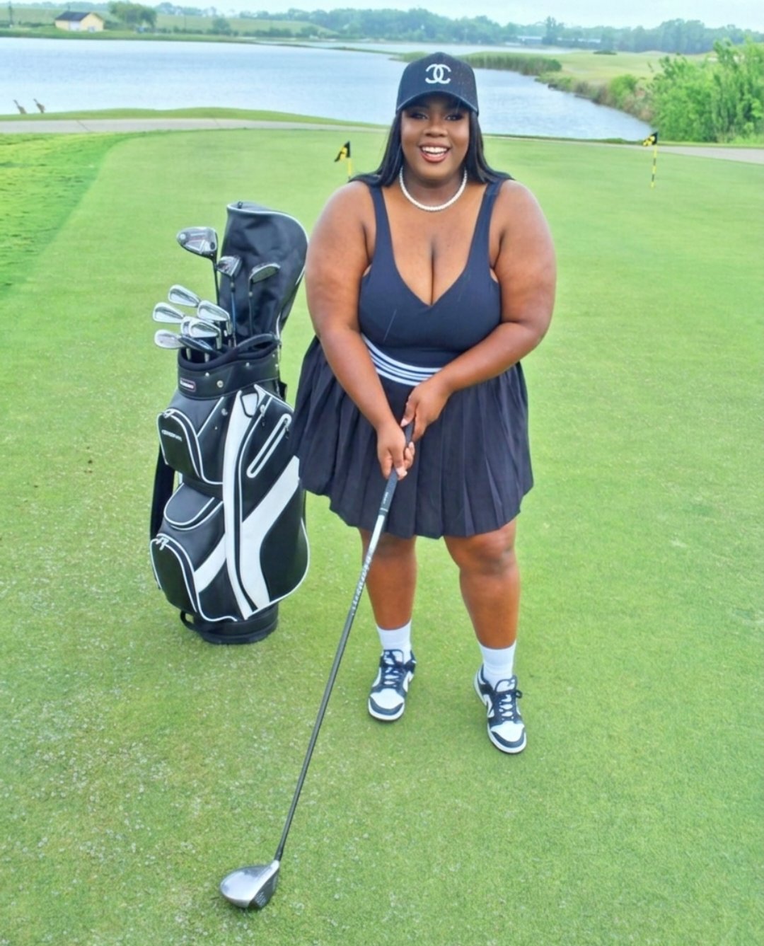 Smiling woman in a navy dress and cap on a golf course, holding a golf club, with a golf bag nearby.