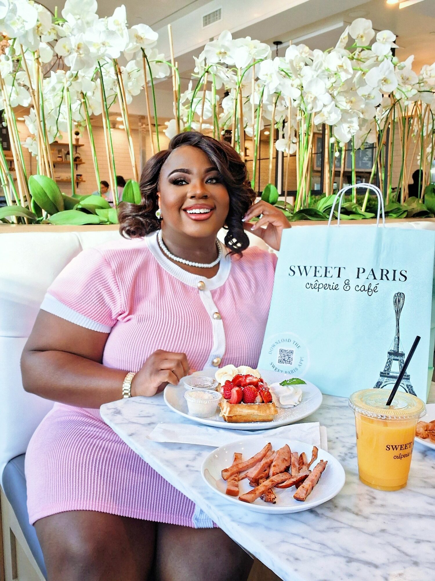 Smiling woman in pink dress sitting at a table with desserts and a shopping bag, surrounded by white flowers.