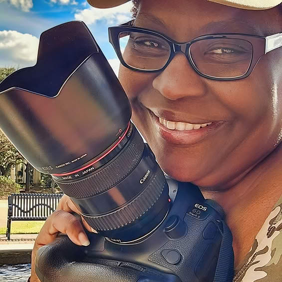 Person smiling, wearing glasses and a hat, holding a camera with a large lens outdoors.