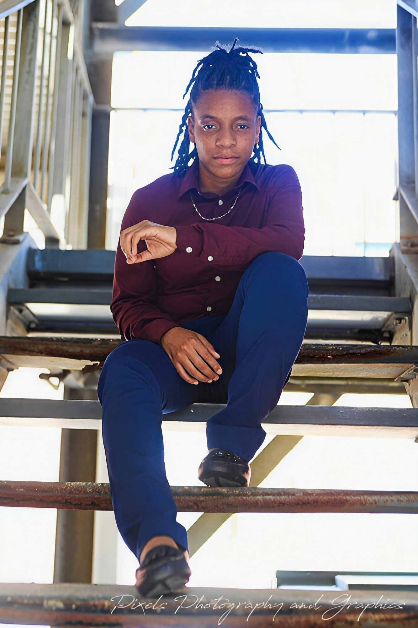 Young woman with dreadlocks sitting on stairs, looking at camera, wearing a maroon shirt and dark pants, in a bright setting.