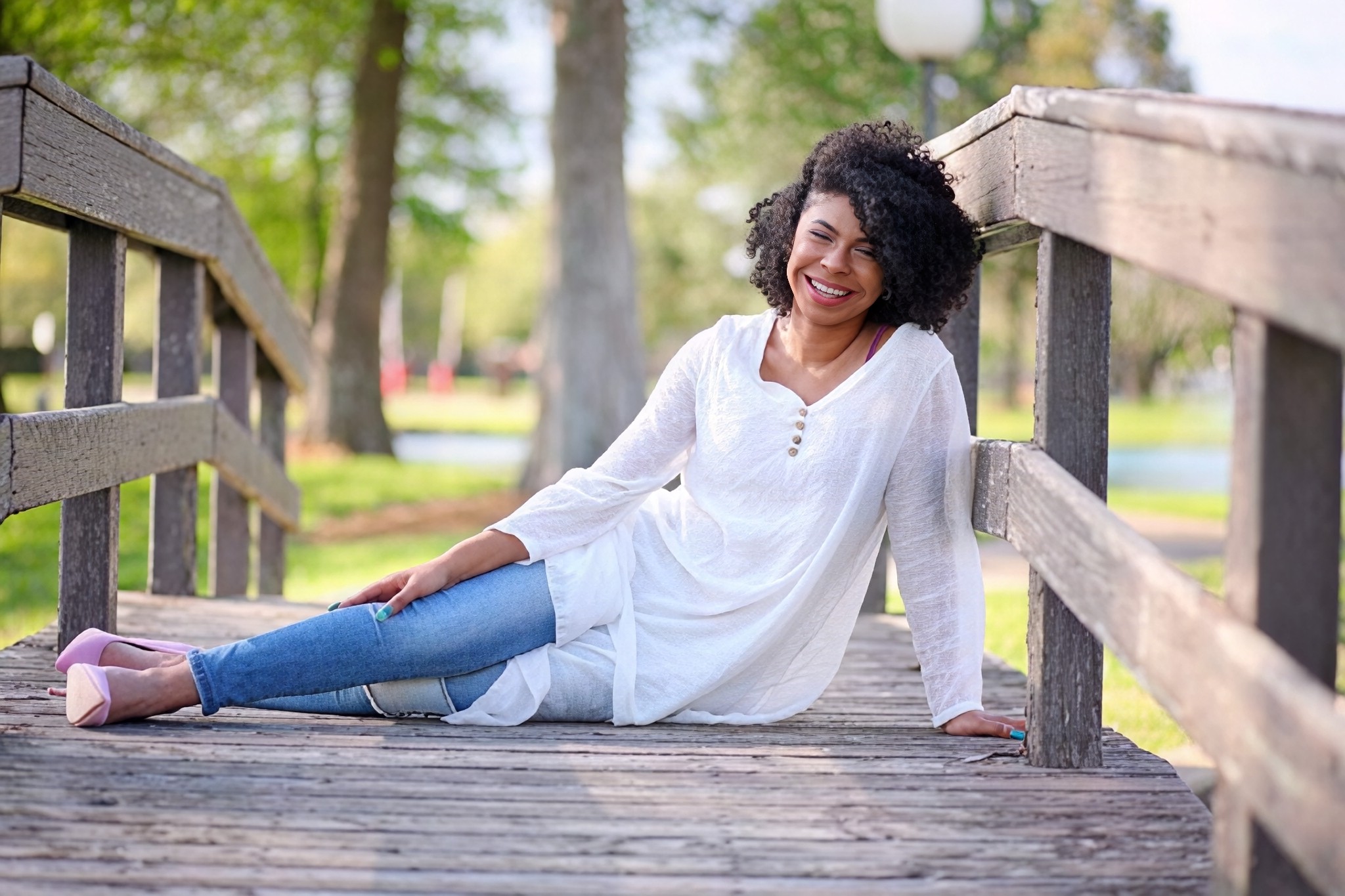 Woman with curly hair sitting on a wooden bridge, smiling, outdoors with trees in the background.