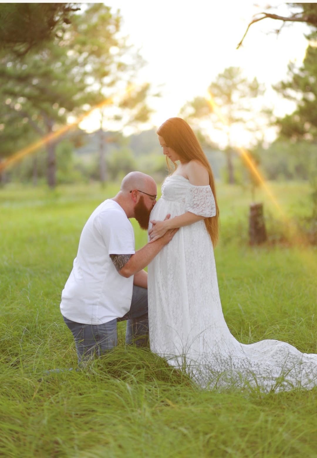 A man kneels holding a woman's hand in a grassy outdoor setting with trees and sunlight, woman in a white dress.
