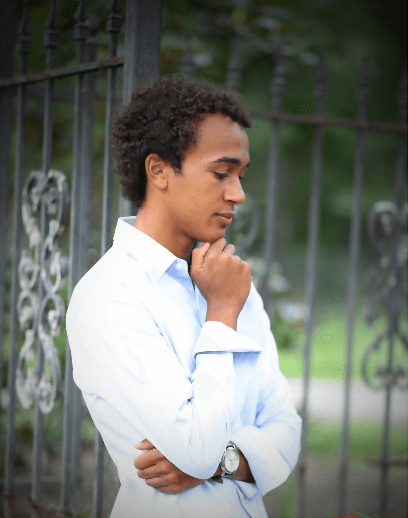 Young man with curly hair in a white shirt, standing outdoors near a metal fence, looking thoughtful with hand on chin.