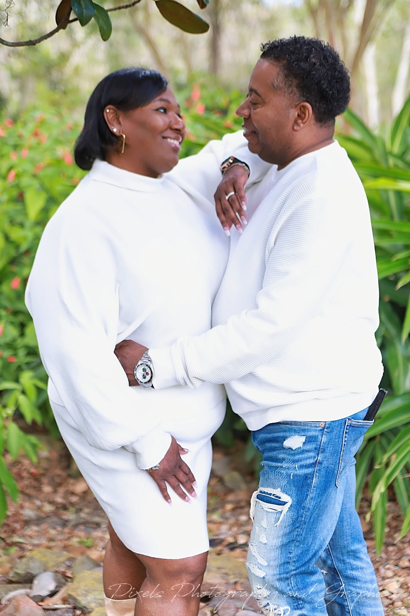 A woman and a man stand close together outdoors, smiling and looking at each other, surrounded by greenery.
