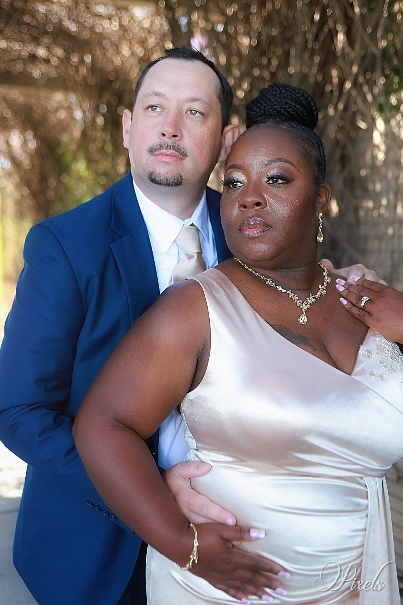 A man and woman pose outdoors, the man in a blue suit and the woman in a white dress, with trees in the background.