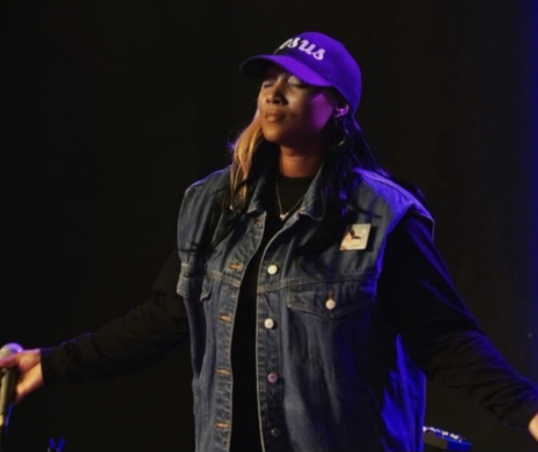 Person wearing a cap and denim vest with arms outstretched, standing on stage with dark background.