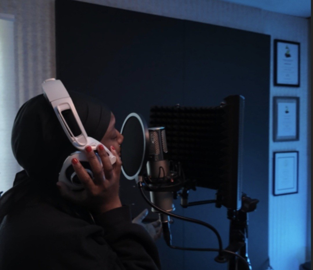 Person wearing headphones in a recording studio with a microphone and soundproofing panels, illuminated by blue light.