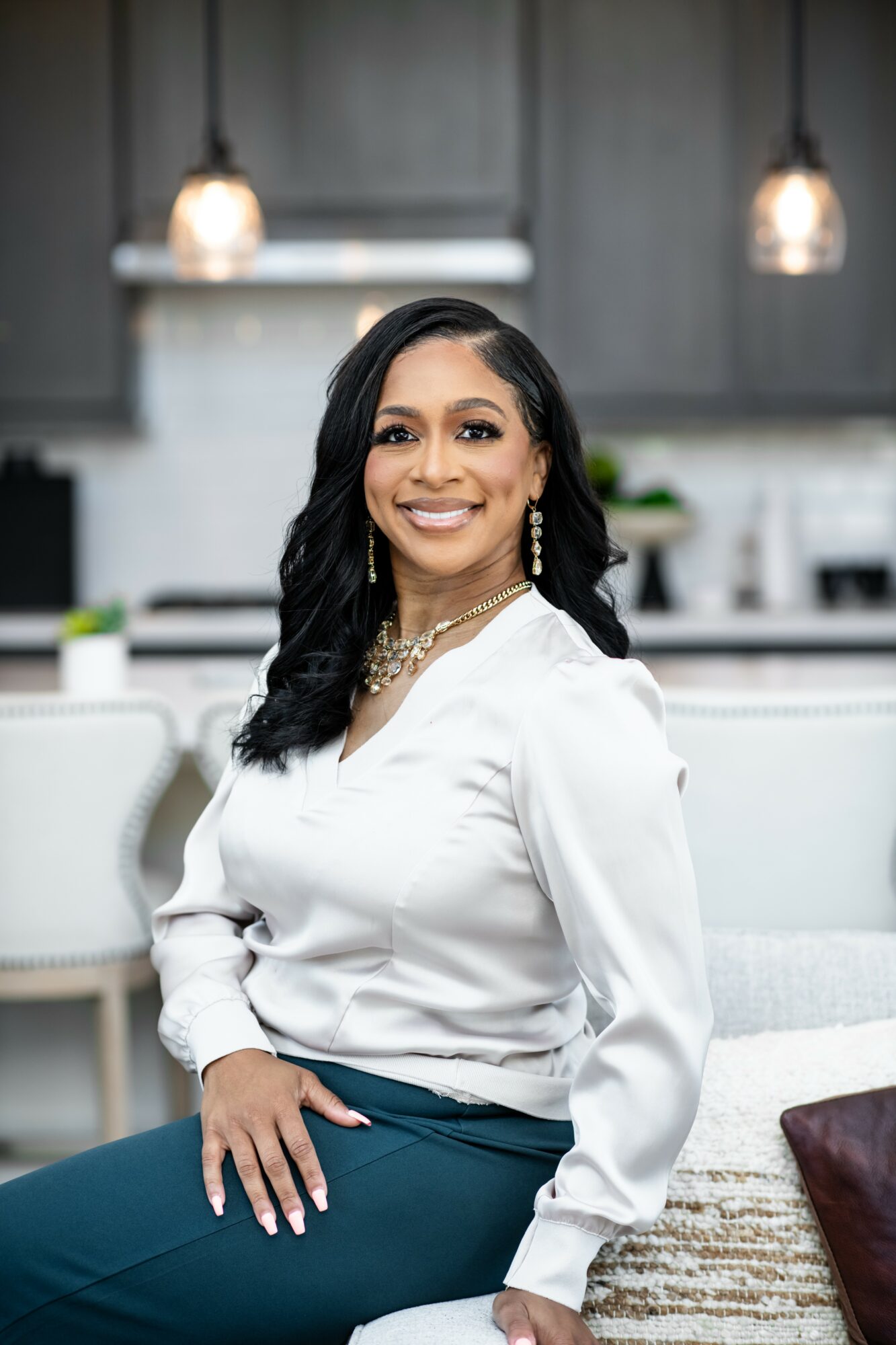 Woman with long black hair smiling, wearing a white blouse and dark pants, sitting in a modern kitchen.