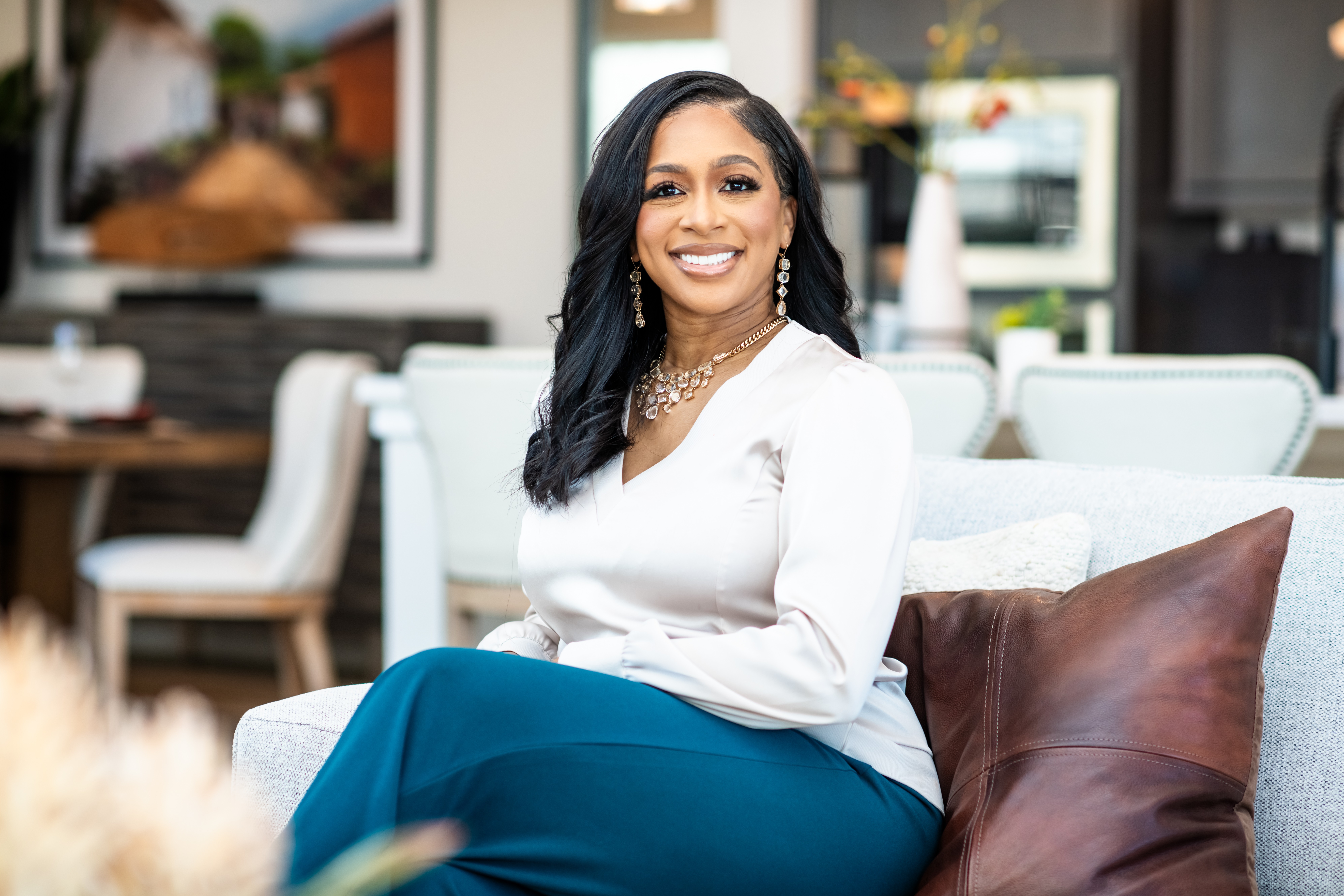 Smiling woman with dark hair and jewelry sitting on a sofa in a bright room.